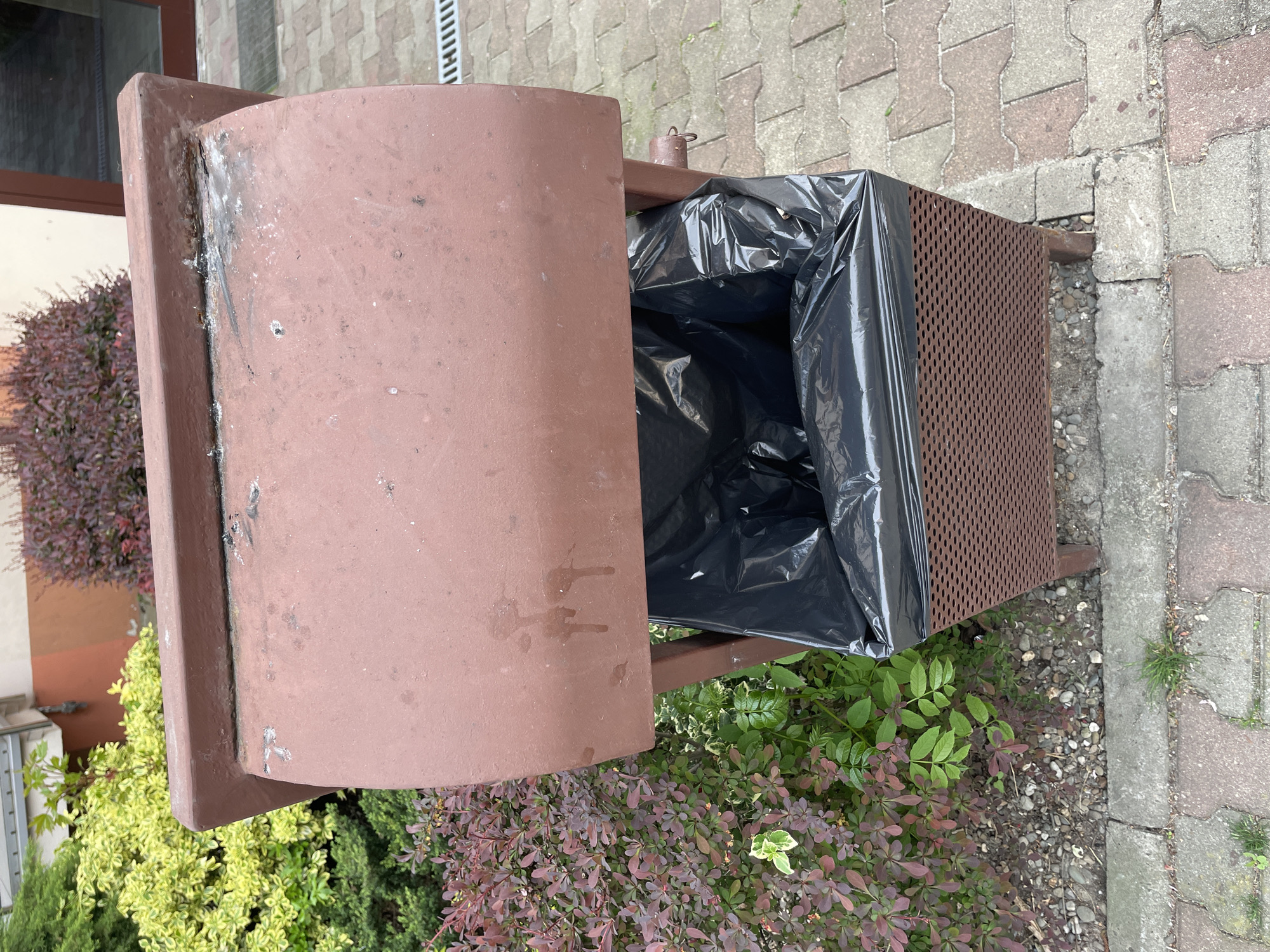 The image shows an open, outdoor trash receptacle with a black plastic bag liner. The receptacle is made of brown metal and appears to be located on a sidewalk or paved area near some greenery and a building.