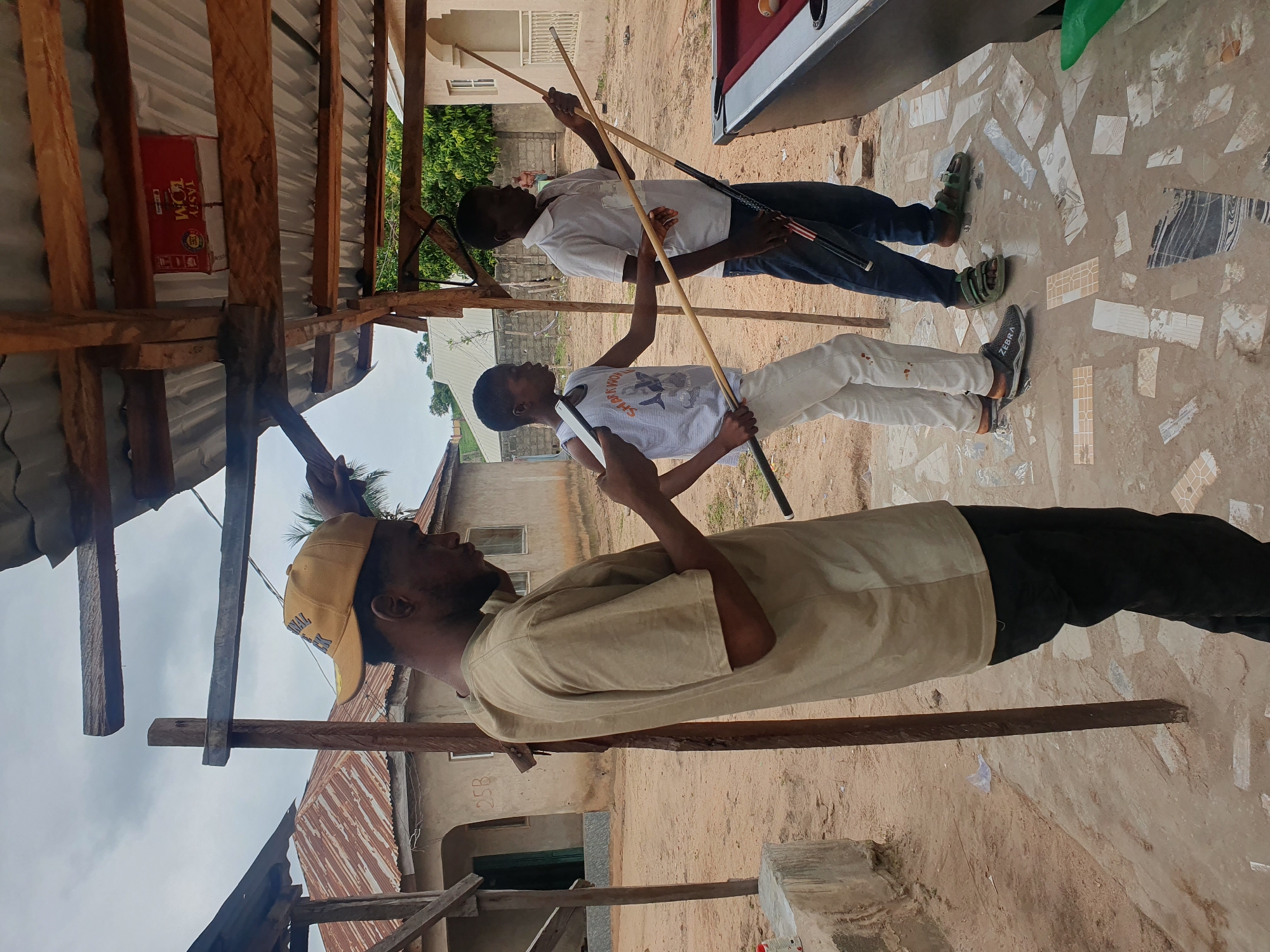 In Lokoja, Nigeria, under a makeshift tin roof structure supported by wooden poles, three young Black males are engaged in a game of pool. The ground is paved with a mosaic of broken tiles and cement. To the left, a male in a tan t-shirt and black pants, wearing a yellow baseball cap, is looking at his phone. In the center, another male in a white tank top and light-colored pants holds a pool cue. To the right, a third male in a white polo shirt and blue jeans, wearing green sandals, also holds a pool cue. A red-topped pool table is visible on the right side of the frame, with a few billiard balls on its surface. In the background, simple buildings with corrugated iron roofs are visible, suggesting a residential area. The sky is overcast.