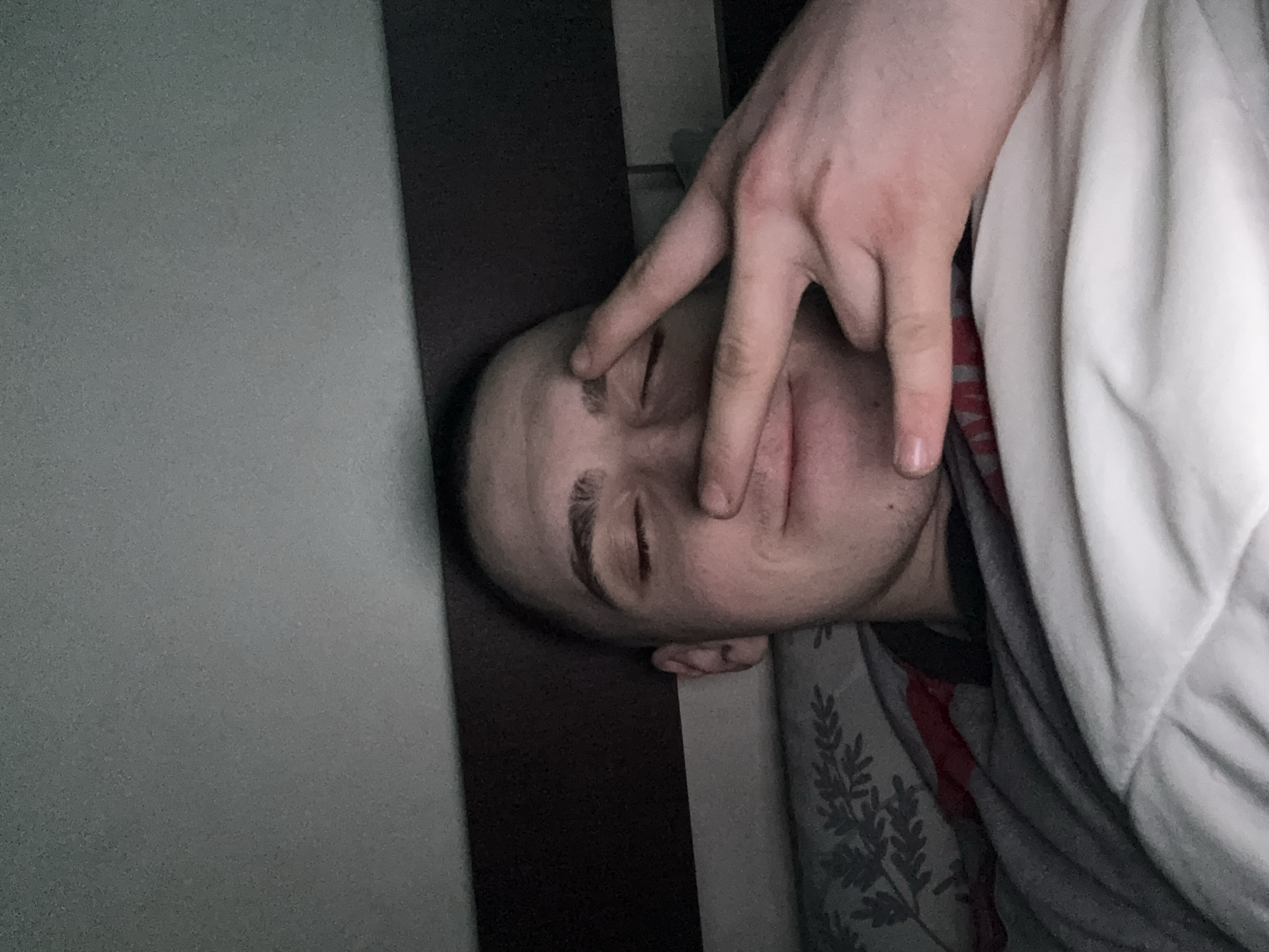 A person is lying down with their eyes closed, and someone's hand is positioned with two fingers extended over their face. The person is partially covered by a white blanket and a patterned gray and red covering. The location is not discernable, and there is no information to conclude the location is Sofia, Bulgaria.