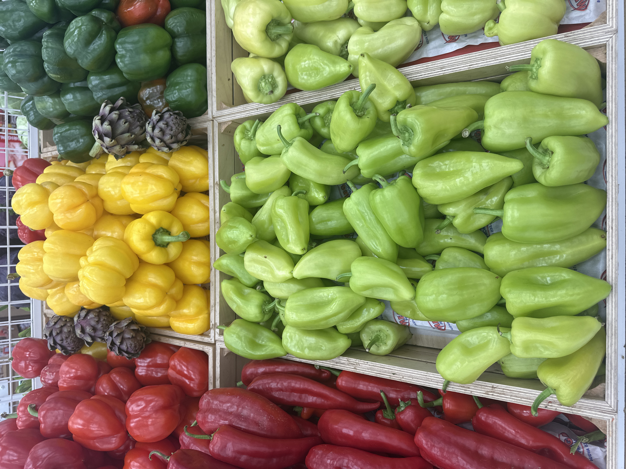 This image shows a display of vegetables in what appears to be a market or grocery store. The image is divided into sections, with each section containing a different type of vegetable.
From left to right, and top to bottom:
1. Green bell peppers.
2. Two artichokes.
3. Yellow bell peppers.
4. Red bell peppers.
5. Light green elongated peppers.
The image was likely taken in Budva, Montenegro.