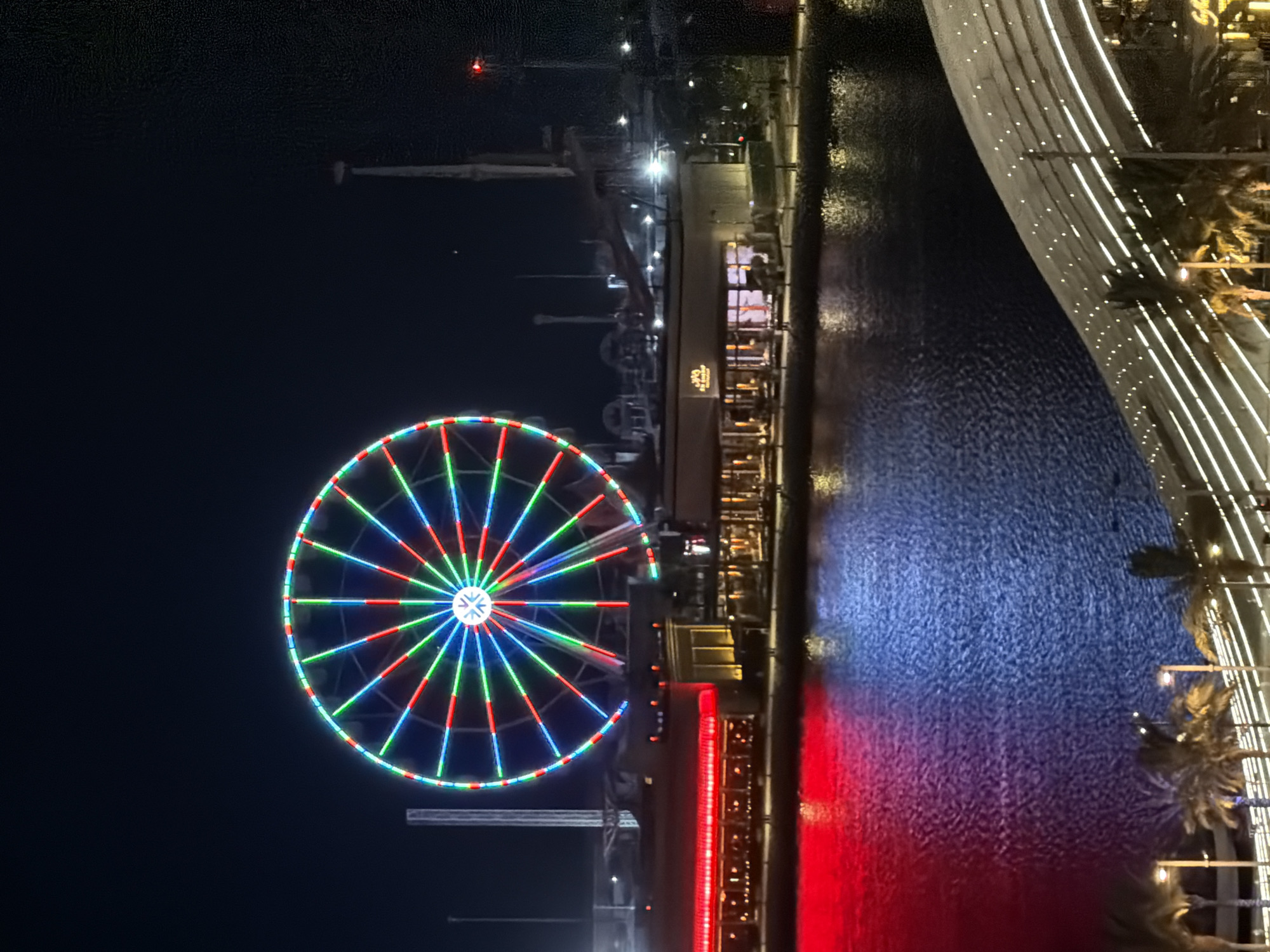 In a nighttime scene, a large Ferris wheel illuminated with multi-colored lights dominates the left side of the frame. The wheel's lights alternate between red, green, and blue, creating a vibrant spectacle against the dark sky. Below the Ferris wheel, a body of water reflects the lights from the Ferris wheel and adjacent buildings. The water is divided into two distinct color zones: a vibrant red on the left and a deep blue on the right.
On the far side of the water, a series of low-rise buildings are visible. These buildings are also illuminated, with some featuring red lighting and others with warm yellow interior lights. One building has a sign that reads "The Hooters". To the right of the Ferris wheel, tall structures that appear to be part of other amusement park rides or attractions are silhouetted against the sky. The overall environment suggests an entertainment district or amusement park at night. In the foreground, a curved structure with white lighting lines suggests a walkway or architectural element. Palm trees are visible in the lower left and right portions of the image, adding a tropical element to the scene.