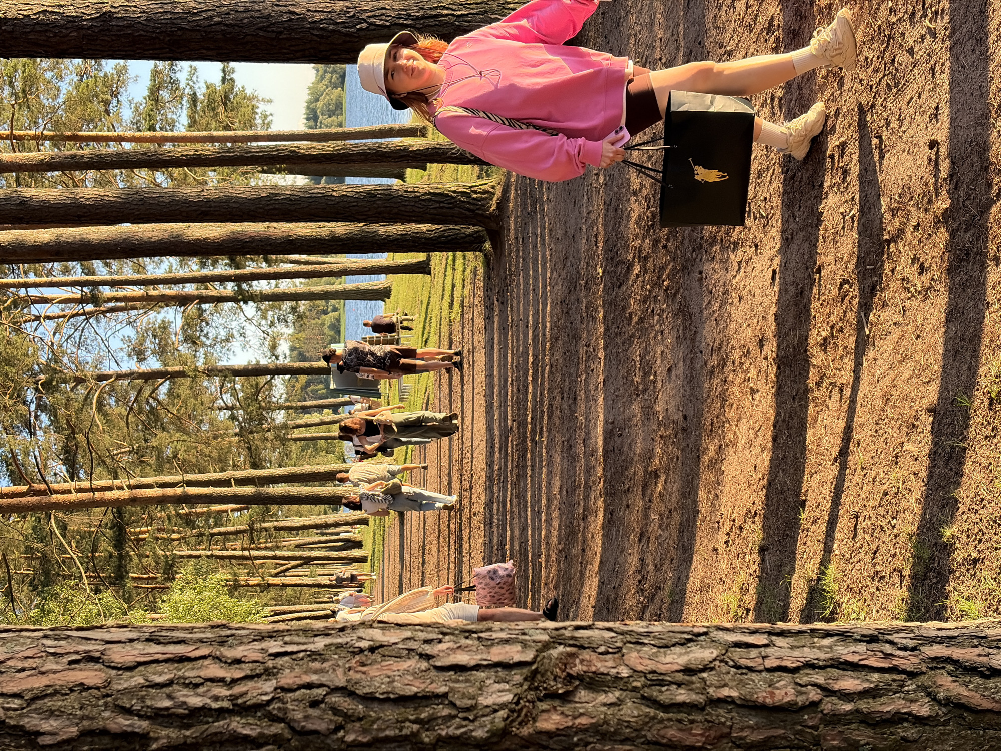 The image depicts a group of people walking along a dirt path lined with tall pine trees. In the foreground, a young woman with reddish-blonde hair is walking towards the right, wearing a pink sweatshirt, dark shorts, white socks, and light-colored sneakers. She is carrying a black shopping bag with a yellow logo. Behind her, several other individuals are walking in the same direction, some carrying bags. The path is dappled with sunlight and shadows from the trees. In the background, a body of water, likely a lake, is visible through the trees, with a tree line and a hazy horizon. The scene appears to be set in an outdoor recreational area, possibly a park or a campground, near a body of water. The environment is characterized by a natural, wooded setting with sandy soil and sparse grass. The presence of the lake and the leisurely activity of the people suggest a summer or late spring day. The location is identified as Zhdanovichi rural council, Belarus.