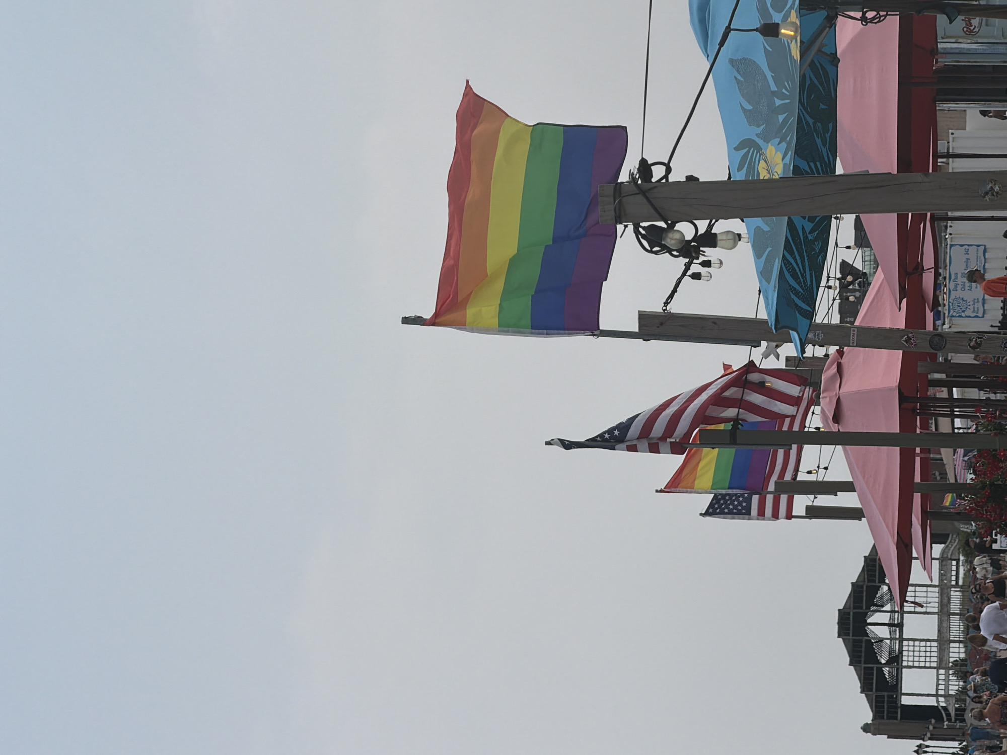 The image depicts a scene in Asbury Park, United States of America, featuring several flags and a crowd of people. In the foreground, a rainbow flag, symbolizing LGBTQ+ pride, is prominently displayed on a flagpole. To the left of the rainbow flag, an American flag is also visible, with parts of its stripes and stars discernible. Further to the left, another American flag is shown, partially obscured.
Behind the flags, a walkway is lined with people dressed in casual attire, suggesting a public gathering or event. Some individuals are walking, while others appear to be standing and conversing. In the background, a large, partially constructed or derelict building with a skeletal frame is visible, characteristic of the Asbury Park boardwalk area. Red and blue umbrellas, likely associated with outdoor seating or vendor stalls, are also present in the scene. The sky is overcast, indicating a cloudy day.