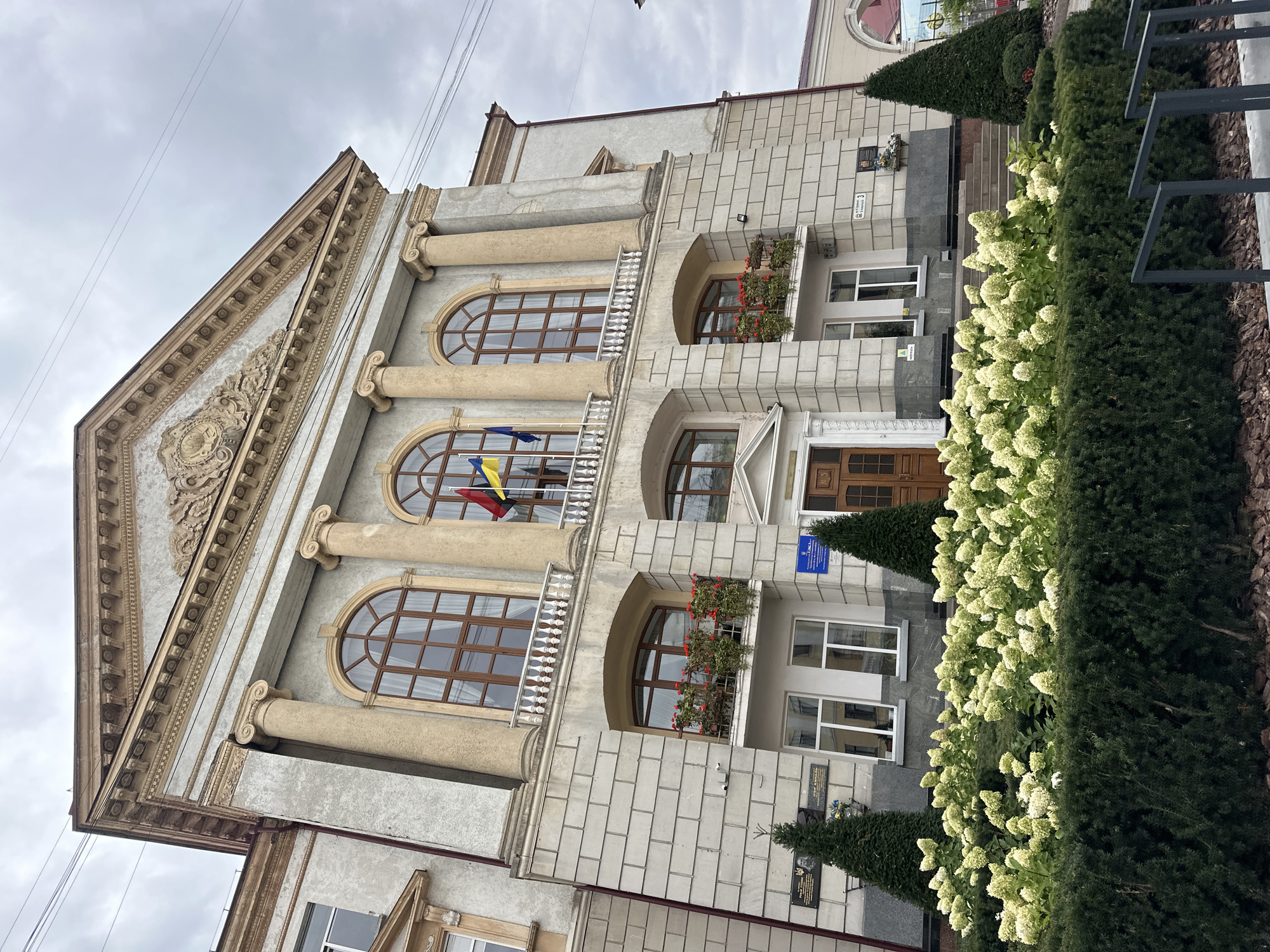 The image shows the Ternopil Regional State Administration building in Ternopil, Ukraine. The building is a grand, multi-story structure with a classical facade, featuring large arched windows, columns, and a triangular pediment adorned with decorative carvings. Ukrainian flags are visible flying from the upper level. In the foreground, there is a well-maintained garden with lush green hedges and a large bed of white flowering hydrangeas. Two conical evergreen trees flank the entrance to the building, and more signage and plaques are visible on the lower facade. The sky is overcast, suggesting a cloudy day.