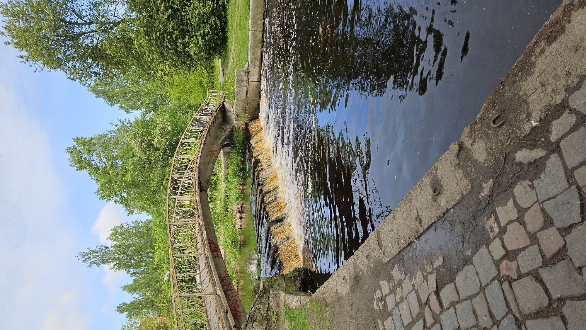 The image shows a waterfront scene in Unknown, Unknown. A small man-made waterfall flows into a body of water, its surface reflecting the sky. A decorative, arched bridge with metal railings spans the water to the left of the falls. Lush green trees line the banks, and a cobbled walkway edges the water on the right.