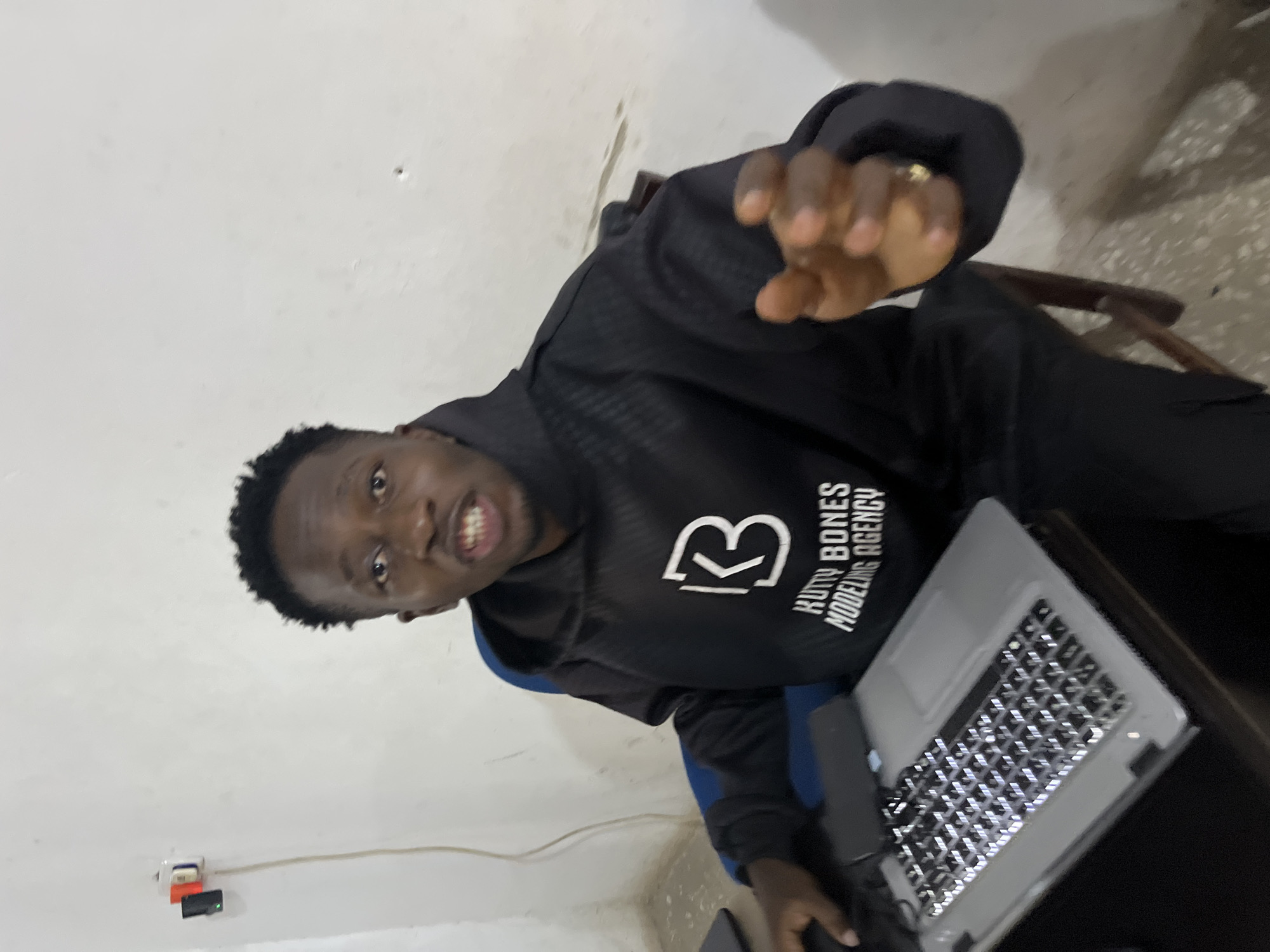 In a sparsely furnished room in Gura Top, Nigeria, a young man with dark skin and short, dark hair sits at a desk in front of a laptop. He is wearing a black hoodie with a white logo that appears to read "KB RUTTY BONES MODELING AGENCY." His left hand is resting on the laptop, which has illuminated keys. His right hand is raised, palm facing forward, as if gesturing or speaking. The room has white walls with some discoloration and a blue chair. Electrical outlets are visible on the wall behind the man, with a power cord extending from one. The overall environment suggests a modest, possibly office-like setting.
