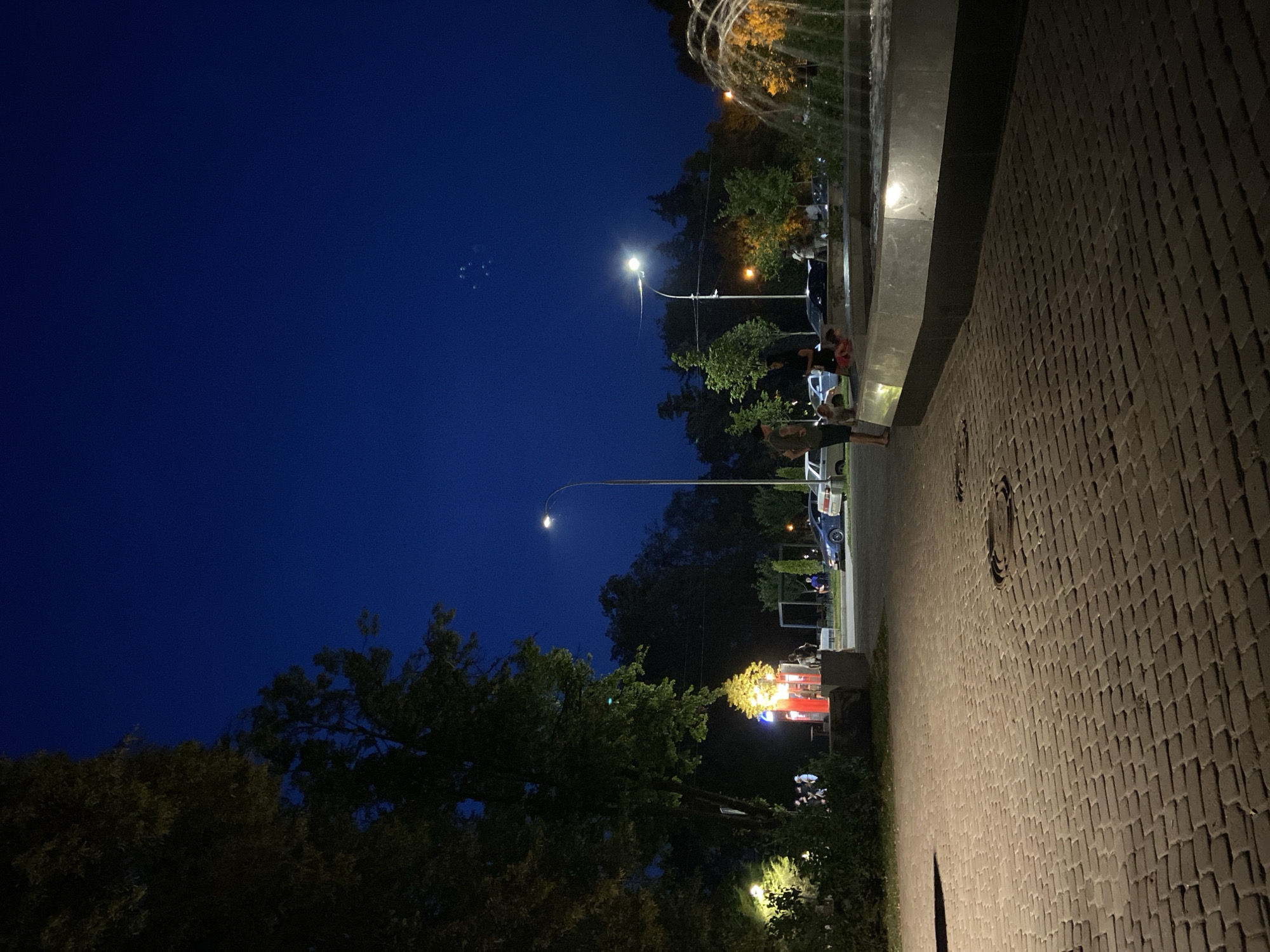 The image depicts a public space at twilight in Poltava, Ukraine. A paved walkway, constructed of brick-like pavers, occupies the foreground, sloping gently upwards and to the right. A fountain with cascading water is visible on the right side of the frame, illuminated from within, reflecting on the wet surfaces of the fountain's basin.
In the mid-ground, a street or open area is visible, lined with trees and streetlights. Several cars are parked along the side of the road. To the left of the walkway, a cluster of people are gathered near a brightly lit structure that appears to be a kiosk or small building, possibly a food stand, with red and blue accents. Further back, more trees and distant lights suggest a park or urban environment.
Several individuals are present in the scene. Near the fountain, a few people are standing or sitting, and one person appears to be interacting with the water. On the walkway, a person is standing near the edge of the fountain. Near the kiosk, a small group of people are congregated, some appearing to be in conversation. Another individual is standing near the kiosk entrance. In the background, more figures are visible, some seated at tables under a canopy.
The sky is a deep, dark blue, indicating the late evening. The overall atmosphere is one of a public gathering space at the end of the day, with artificial lights illuminating the scene against the darkening sky.