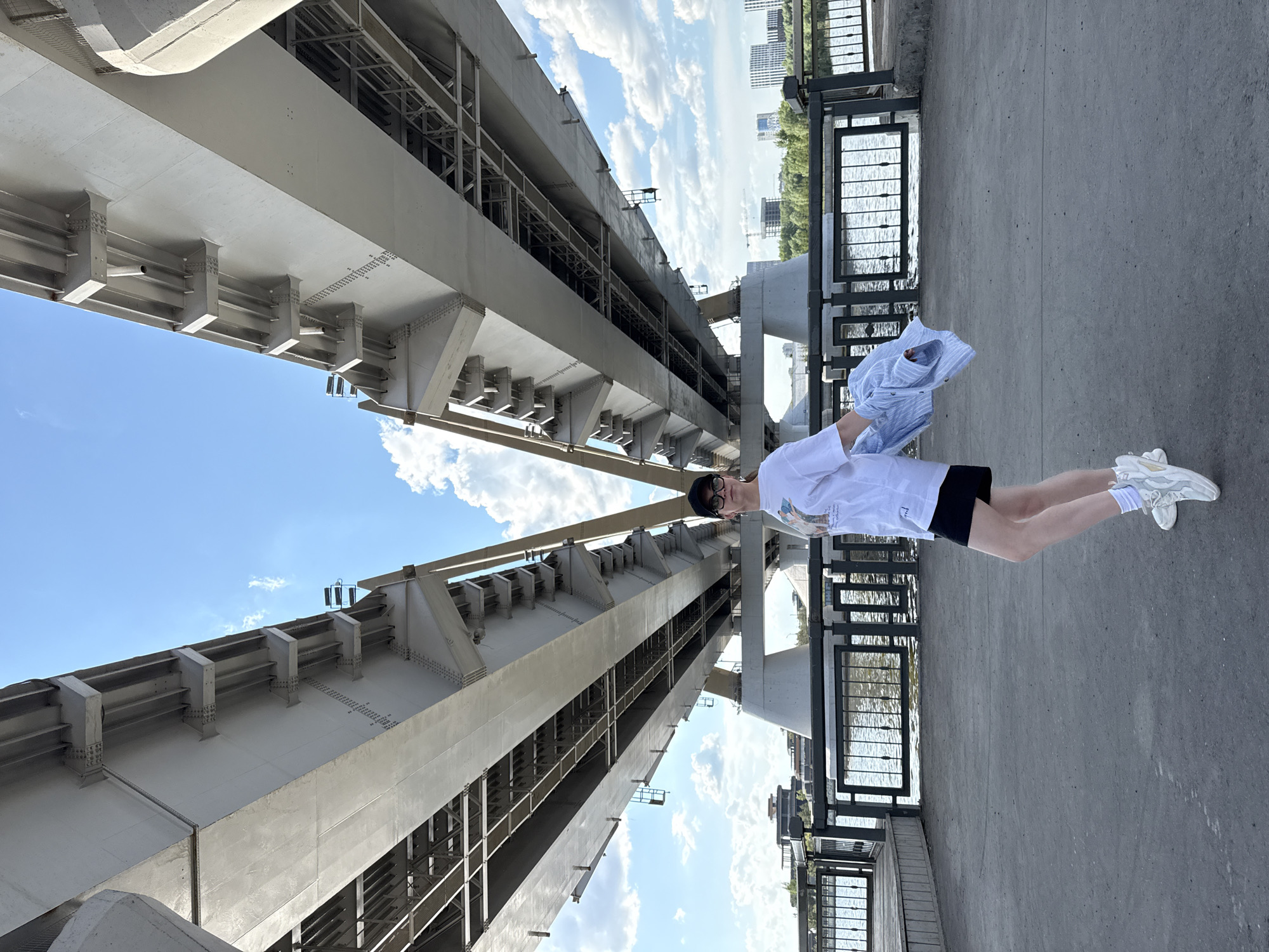 A young person stands on a concrete surface next to a metal railing, holding a blue striped shirt. The person is wearing a white T-shirt, black shorts, white socks, and white sneakers. The background features the framework of a bridge. Beyond the railing, there is a body of water and a distant urban skyline. The scene appears to be photographed from an unusual angle, as if the camera is tilted to emphasize the height of the bridge structure. Based on the bridge structure and urban context, the location is likely in Kazan, Russia.