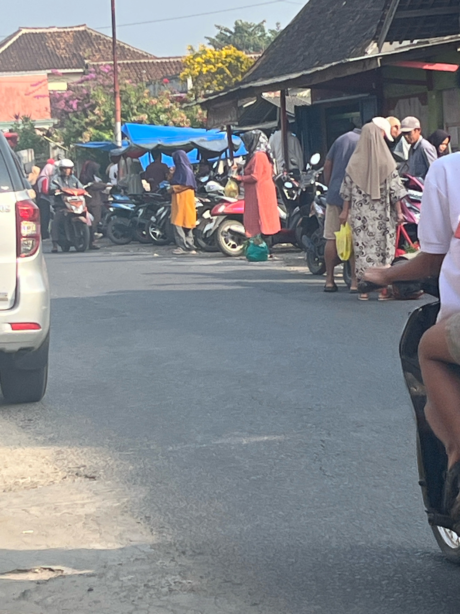 A street scene in Kemuning, Indonesia, captures a bustling market area. Several individuals, some wearing hijabs, are gathered around parked motorcycles and makeshift stalls. One person, wearing an orange garment, stands near a motorcycle. Another individual in a floral patterned dress and beige hijab is visible. A silver SUV is parked on the left side of the road. The background shows buildings with tiled roofs and some greenery. The pavement is rough and shows signs of wear.