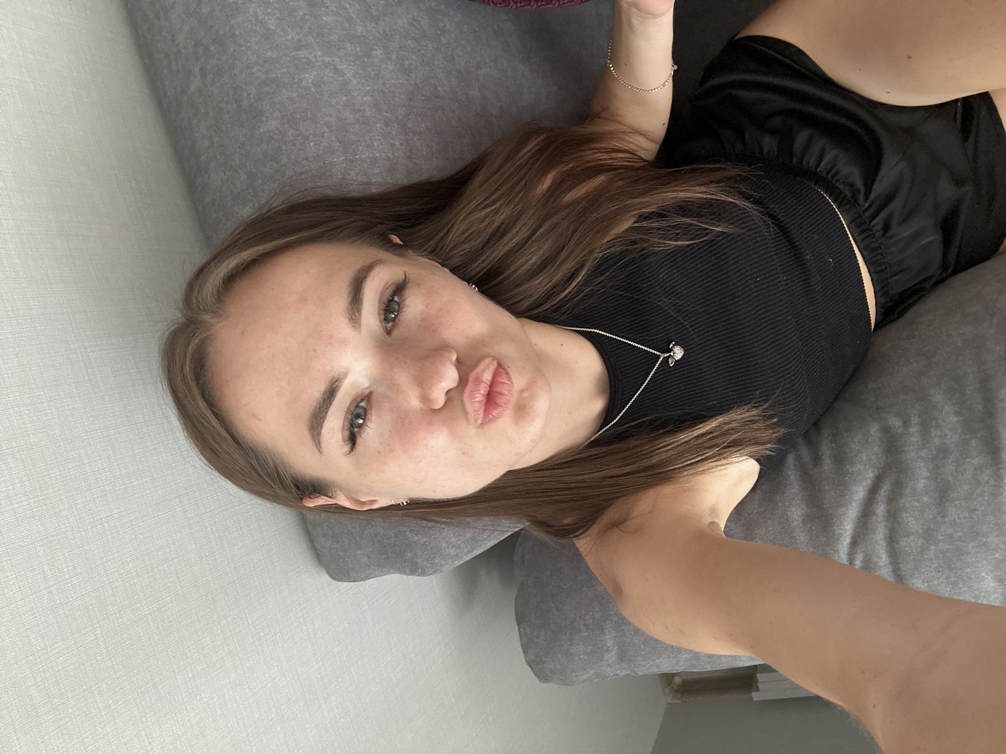 A young woman with long, brown hair is lying on a gray sofa, posing for a selfie. She is wearing a black ribbed tank top and black silk shorts. A silver necklace with a heart-shaped pendant is visible around her neck, and she is wearing a delicate bracelet on her wrist. Her right arm is extended, holding the camera. The background consists of the textured gray fabric of the sofa and a light-colored wall with a subtle textured pattern.