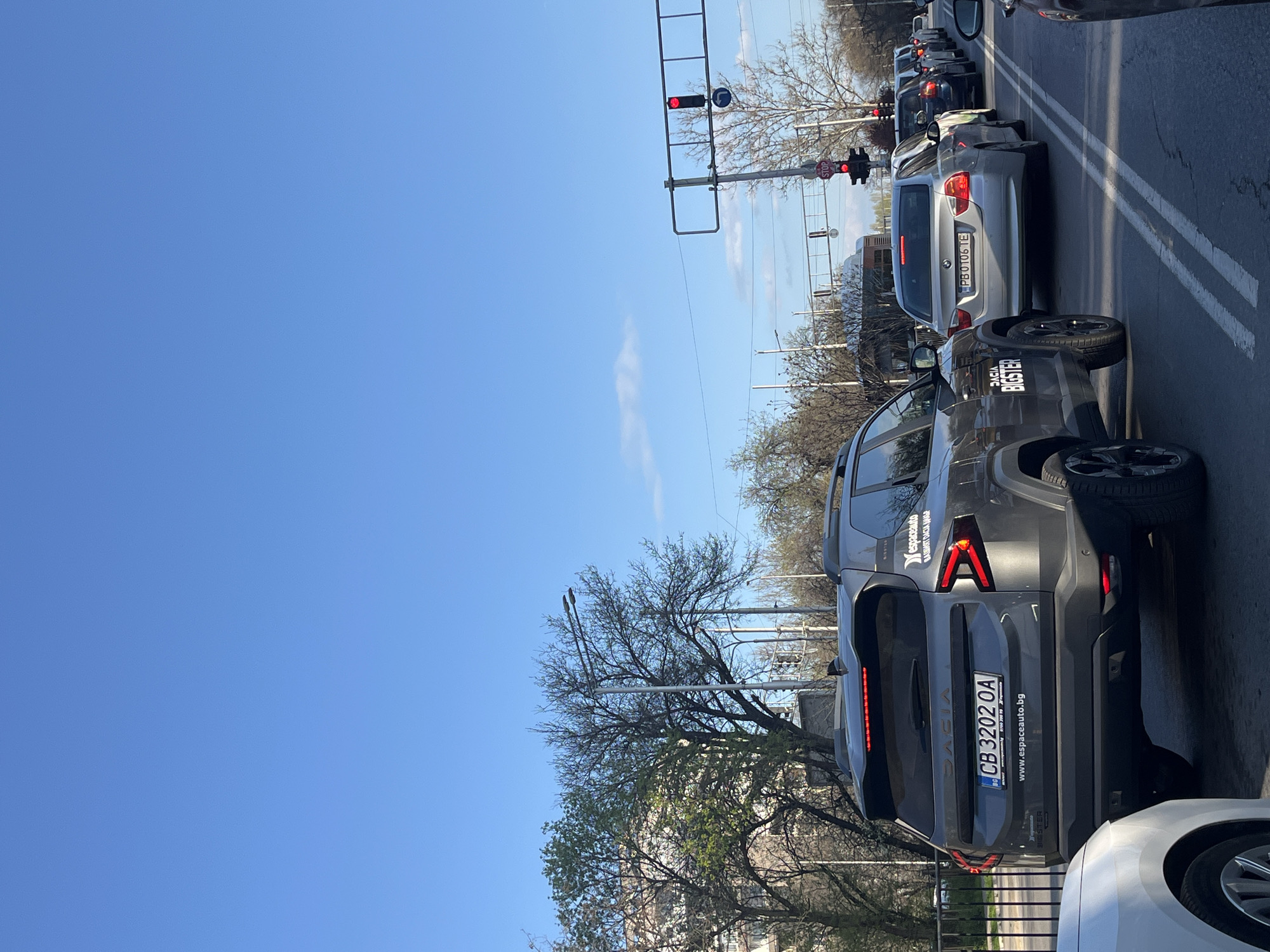 The image shows a street scene in Plovdiv, Bulgaria. Cars are stopped at a traffic light, where the light is red. A gray Dacia Bigster is in the foreground, with the license plate CB 3202 OA, affiliated with "espaceauto.bg". Behind the Dacia is a silver BMW with license plate PB 0106 TE. In the background, several other vehicles are lined up behind the stopped cars, waiting for the light to change. The sky is clear and blue. Trees line the street.