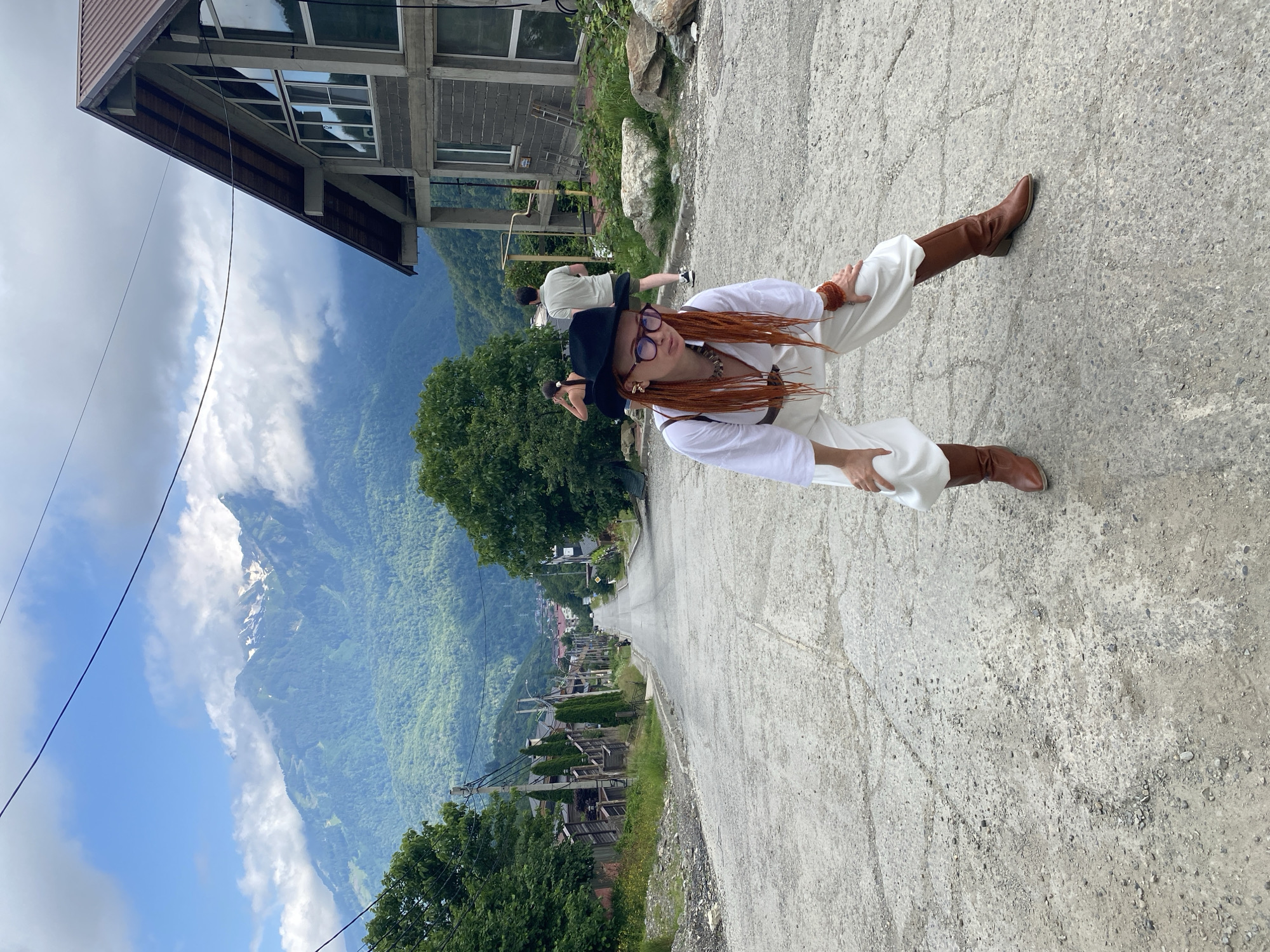The image depicts a woman with bright orange braided hair and a black hat, wearing a white shirt and loose white pants, and brown boots. She is in a squatting pose on a paved road that slopes downwards into a valley. In the background, there are mountains, trees, and buildings, including a distinctive modern house with a large glass facade. The scene appears to be located in Krasnaya Polyana, Russia, a mountain resort town known for its ski slopes and natural beauty, as suggested by the mountainous terrain and the architecture that is typical of such locations. The sky is partly cloudy, with some sunlight breaking through. There are other people visible in the background, including a man and a woman walking away from the camera. The overall atmosphere is casual and outdoorsy.