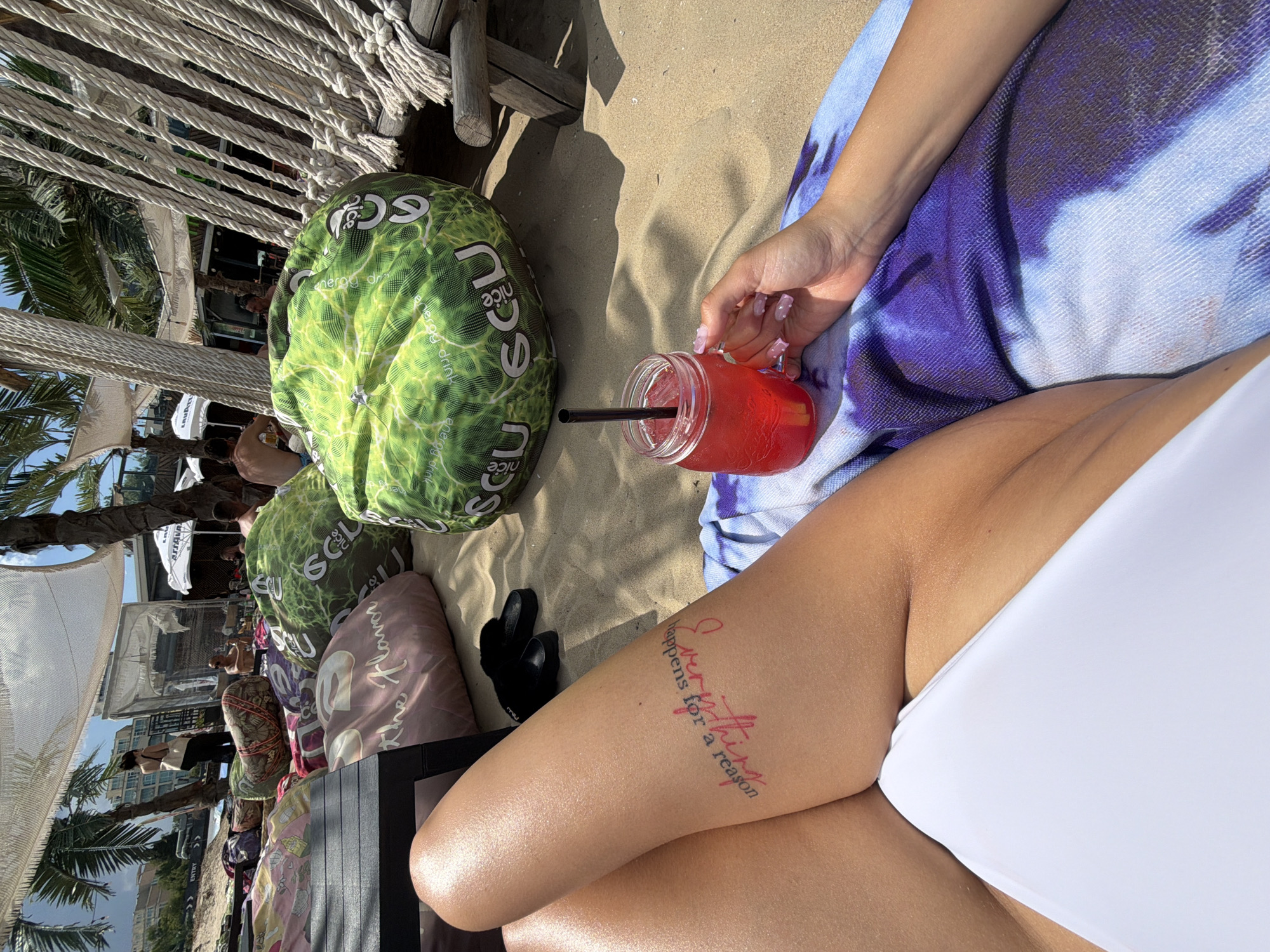 An overhead, close-up shot shows a person's legs and a drink on a beach in Nesebar, Bulgaria. The person is lying down on a purple and white tie-dye towel. They are wearing a white bikini bottom. Their left leg has a pink tattoo that says "Everything happens for a reason." In their right hand, they are holding a glass jar with a red drink and a black straw. In the background, there are large green and pink beanbag chairs and palm trees. The scene is set on a sandy beach with the ocean not visible.
