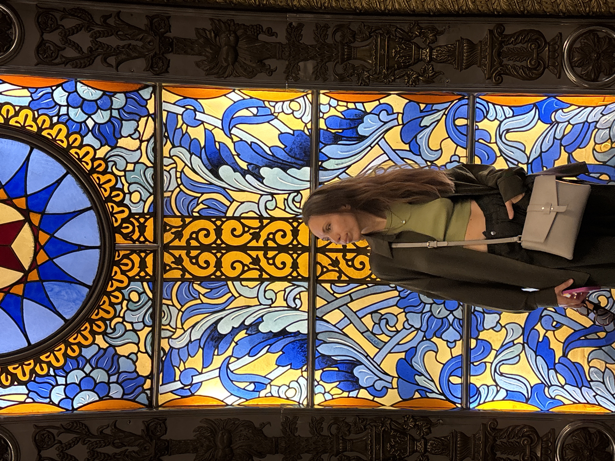 A woman is standing in front of a large, ornate stained-glass ceiling. The ceiling is divided into rectangular panels and a circular section on the left. The panels feature a repeating pattern of blue, yellow, and light blue floral and swirling designs. The circular section has a starburst pattern in blue, yellow, and red. A decorative, dark-colored metalwork border frames the stained glass. The woman is wearing a dark jacket, a light green top, and dark pants. She has long brown hair and is carrying a light-colored bag. She appears to be looking at her phone, which she is holding in her right hand. The setting appears to be indoors, likely within an ornate building.
Given the architectural style and based on the image, the location is likely within the Moscow Metro, specifically a station with notable architectural features, such as Komsomolskaya station.
