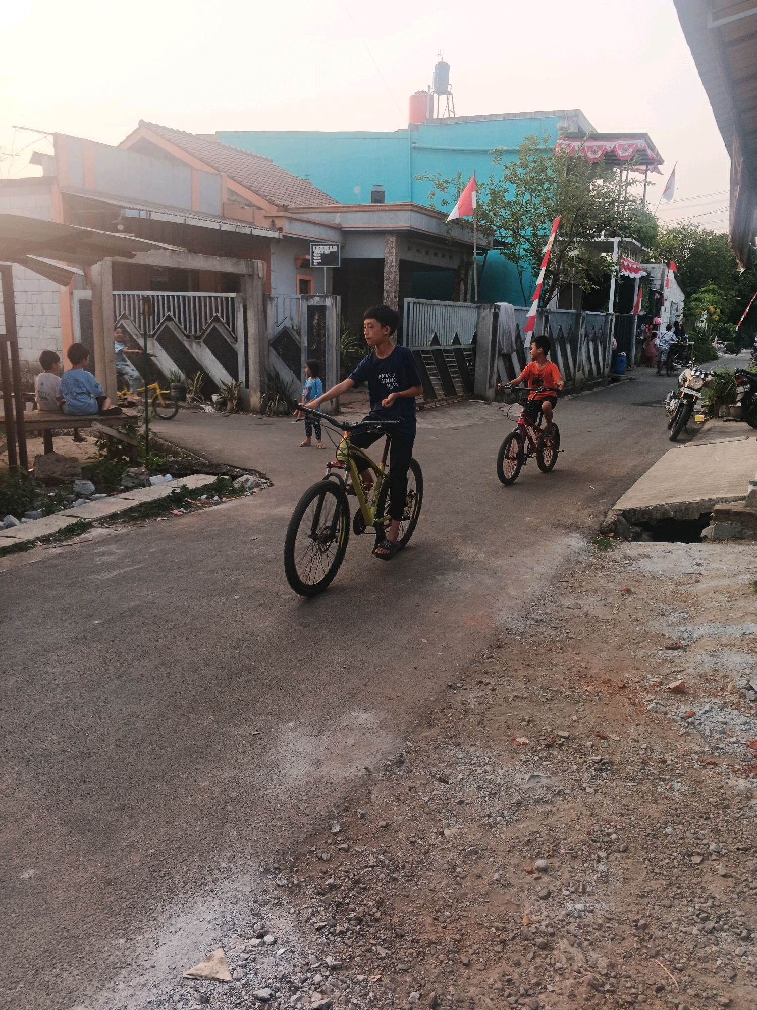 The image captures a street scene in Kadu, Indonesia, under a hazy sky. In the foreground, a young boy on a yellow bicycle is visible, his face turned to the left. Further down the street, another young boy rides a red bicycle, facing forward. The road is asphalt, with a gravelly shoulder. Buildings with tiled roofs line the street, some painted in teal and others in muted tones. Indonesian flags are displayed on some of the buildings, suggesting a celebration or national observance. Several people are seen in the background, and motorcycles are parked on the side of the road. The overall impression is of a quiet residential street, possibly in the late afternoon or early morning given the soft light.