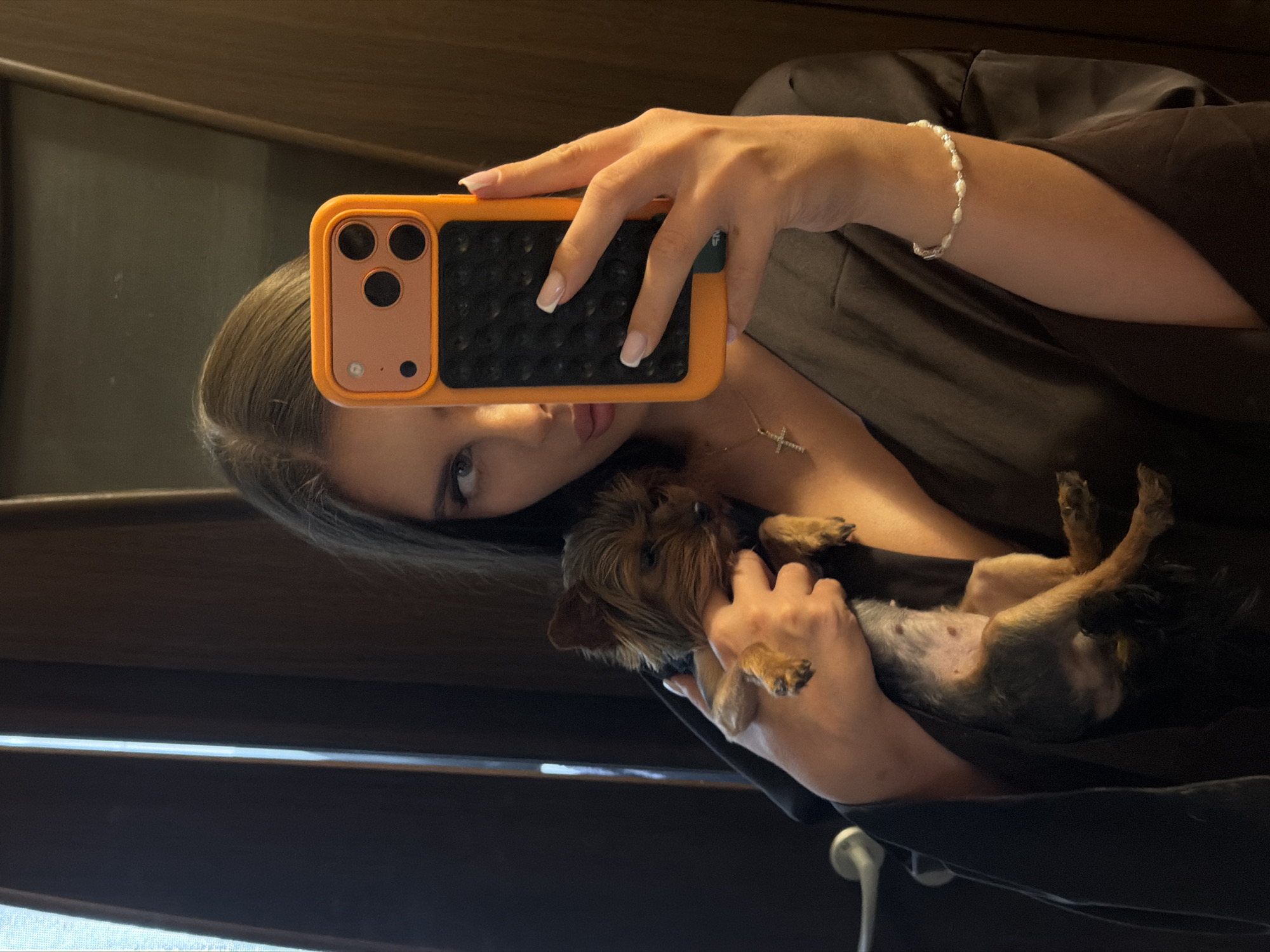 A woman is taking a selfie in a mirror. She is holding a small, brown dog. She has long dark hair and is wearing a brown robe. She is holding an orange phone in front of her face. She is also wearing a silver bracelet and necklace. The background is a wall. There are no identifying details to determine the location is Krasnoyarsk, Russia.