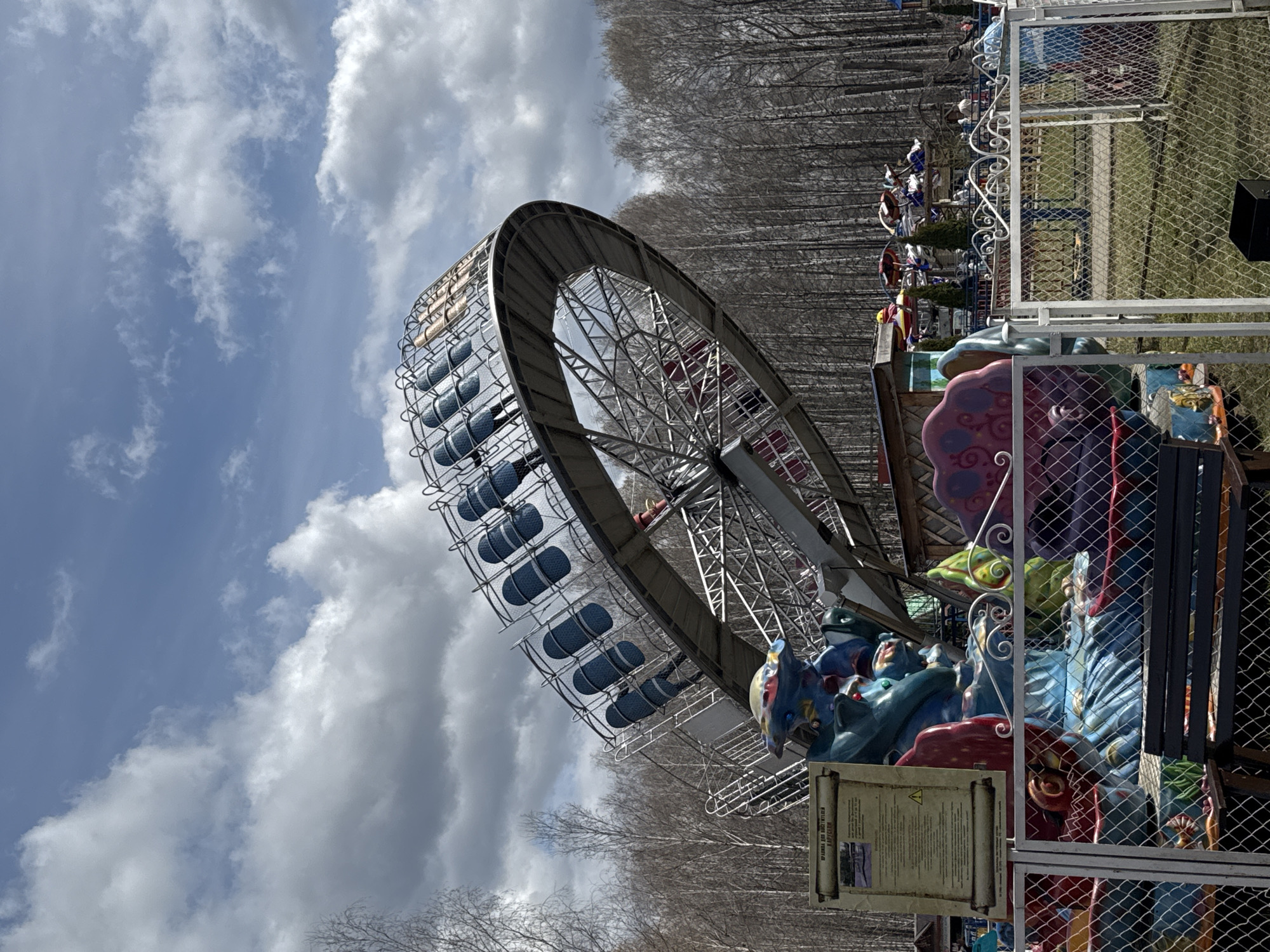 The image shows a fairground ride in the city of Chaikovsky, Russia. The ride has a large circular frame with seats hanging from it. The seats are blue and cylindrical. The frame is made of metal and has a lattice structure. There are bare trees in the background. The sky is blue with white clouds. There is fencing and decorative figures near the ride. A sign with text in Russian is attached to the base of the ride.