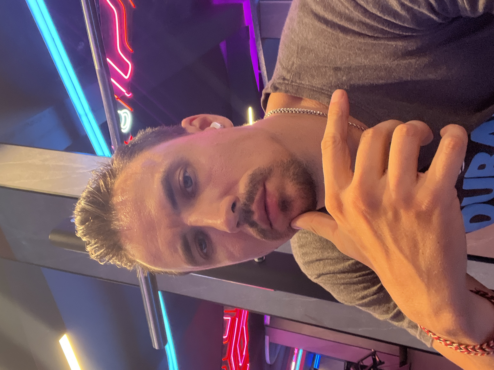 An indoor selfie is taken by a Caucasian man with light skin, short styled brown hair, and a thin mustache and goatee. He is wearing a gray t-shirt, a silver chain necklace, and a white earbud in his right ear. His right hand is resting on his chin, and his left arm, wearing a red and white braided bracelet, is extended towards the camera. The background is dimly lit with neon lights, including blue and pink accents, suggesting an entertainment venue or club in North Kuta, Indonesia.