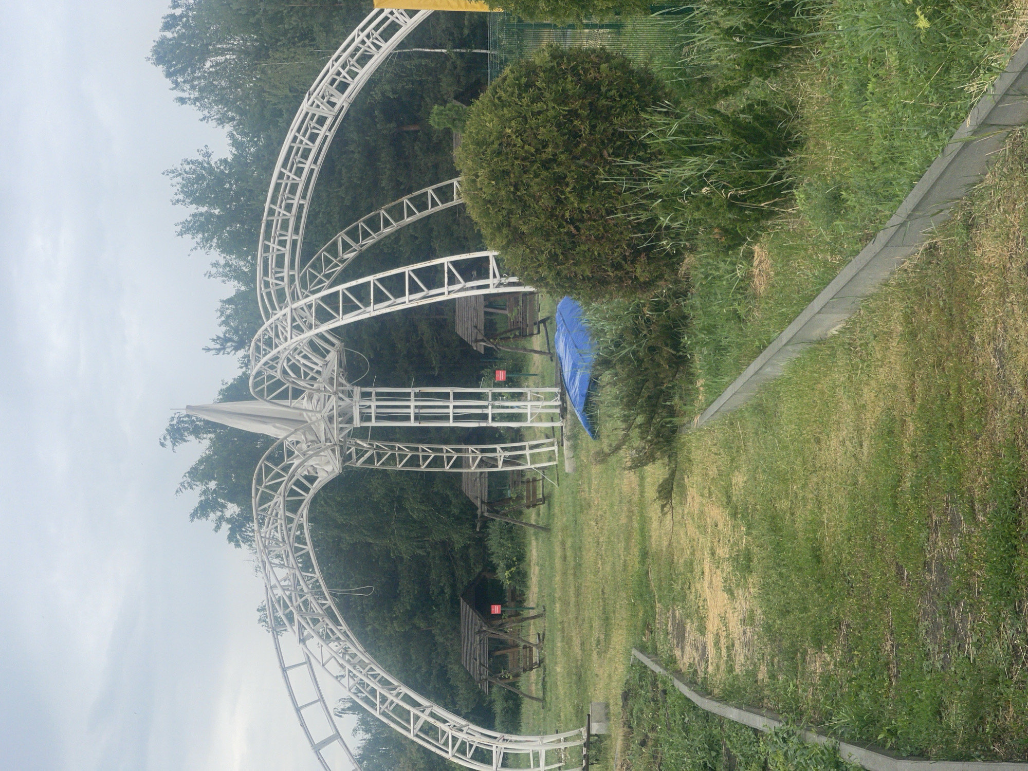 The image depicts an outdoor scene with a large, white, geodesic-like structure in the foreground, possibly a canopy or a decorative archway. The structure is made of interconnected beams and has a fabric covering at its apex. Beneath the structure, there is a grassy area with two wooden swings, each under a small wooden roof. A blue tarp is visible lying on the ground near one of the swings.
To the right of the main structure, there is a large, round, evergreen bush. A green metal fence runs along the right edge of the image, partially obscured by vegetation. A path made of gravel and grass leads into the scene from the bottom left corner, bordered by concrete edging. The background consists of a dense line of green trees, suggesting a wooded area or park. The sky is overcast and appears cloudy. The image is taken from a slightly low angle, looking upwards towards the structure and the trees.