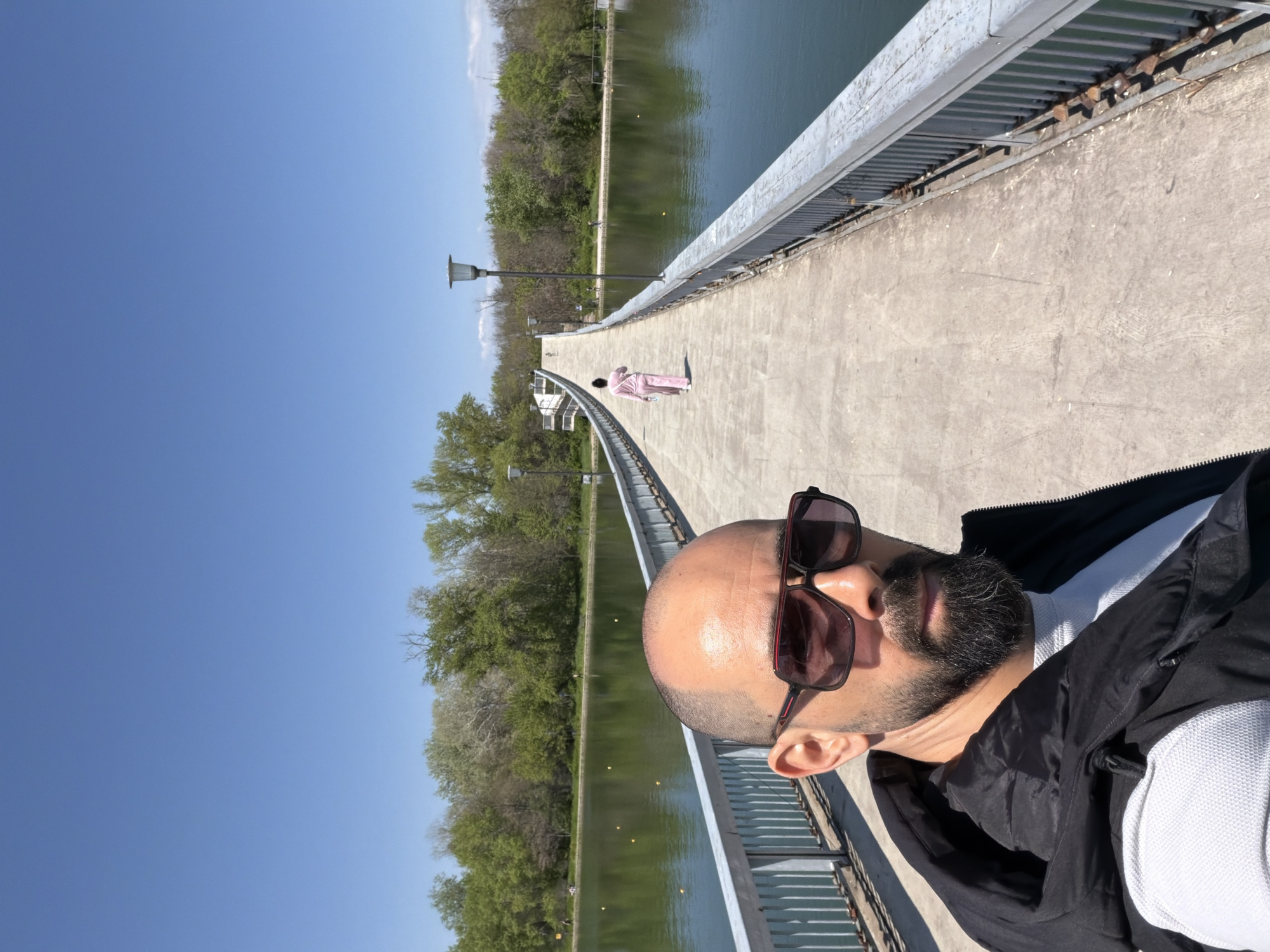 A man wearing sunglasses and a black jacket takes a selfie on a bridge over a body of water, with trees on either side. In the distance, another person in a pink robe walks on the bridge. The location is likely Plovdiv, Bulgaria.