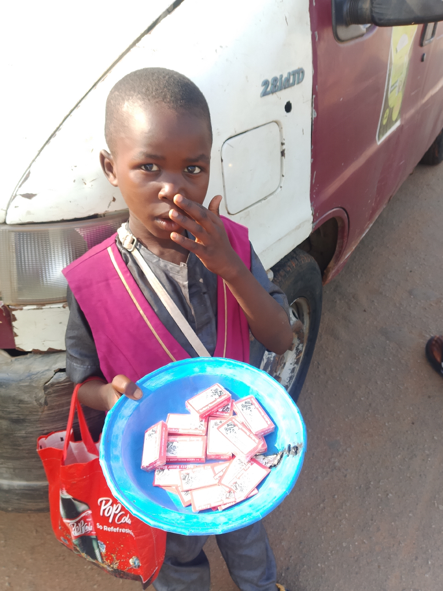 A young boy stands next to a maroon and white van, holding a blue bowl filled with small, red-and-white boxes. He wears a dark shirt with a magenta vest over it and has his right hand near his mouth. A red bag with a "Pop Cola" logo is visible on his left. The van has "2.8D TD" badging on its side. The ground is paved with loose gravel.