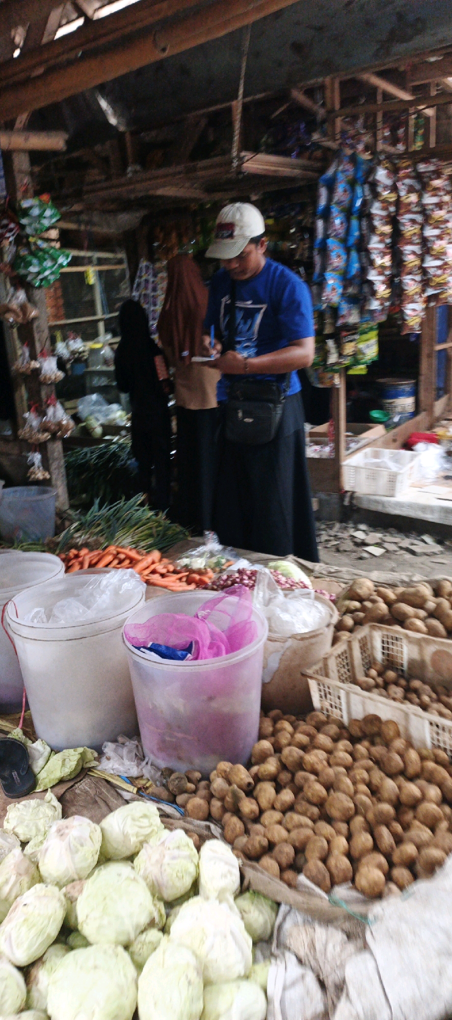 The image depicts a market scene in Kasiyan, Indonesia. In the foreground, a variety of fresh produce is displayed, including piles of potatoes, heads of cabbage, carrots, and onions. Two large plastic buckets, one white and one translucent pink, are visible. A man wearing a blue shirt, a light-colored baseball cap, and a dark cross-body bag is standing in the middle ground, appearing to write on a notepad. To his left, a woman in a brown hijab and a dark long skirt stands beside another woman in a black hijab. The background shows a rustic stall structure with shelves stacked with packaged goods and other market items. The overall environment suggests a bustling local market.