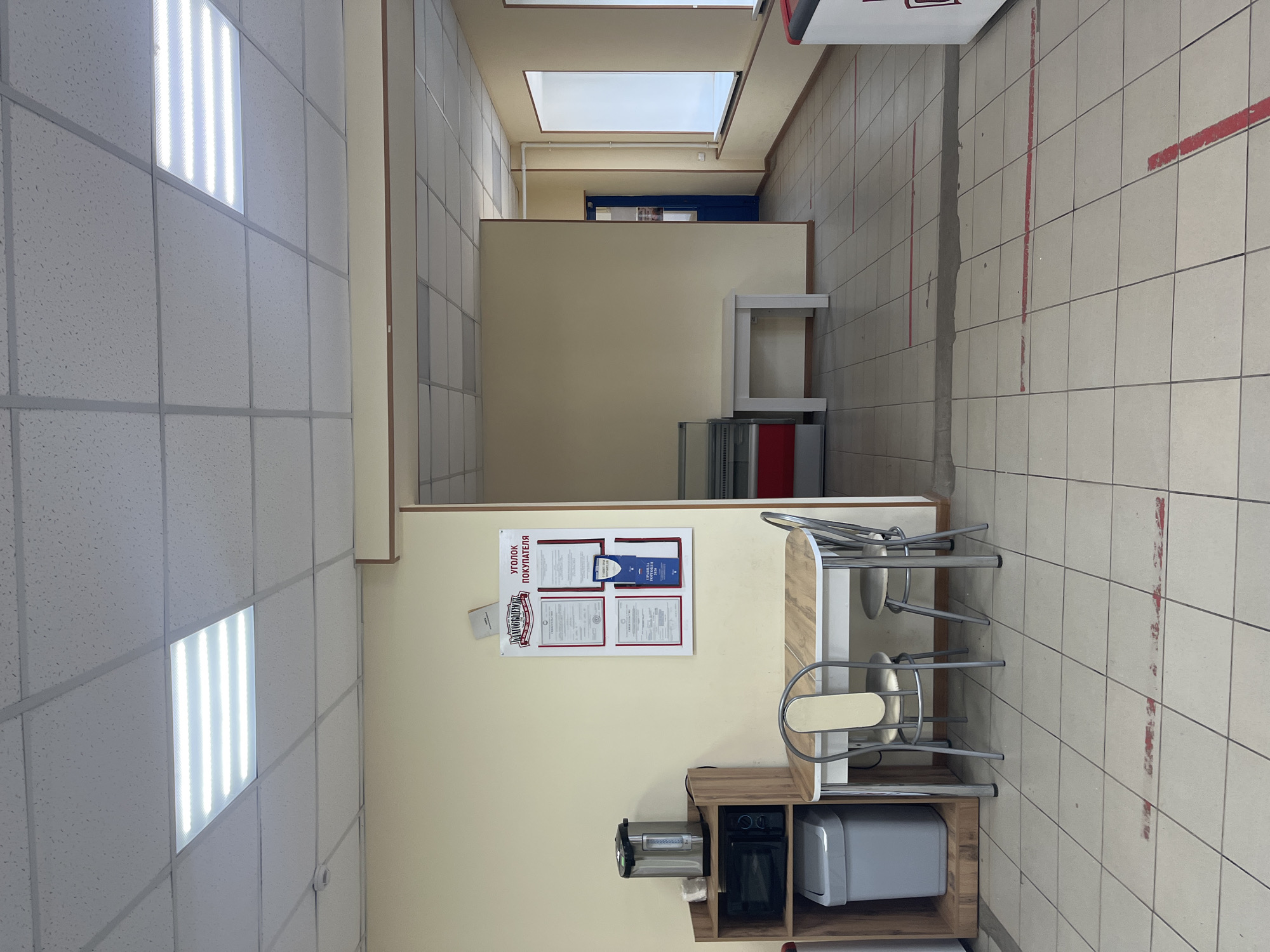 This is an indoor scene, possibly a cafeteria or break room, in Blagoveshchensk, Russia. The space has a tiled floor and a suspended ceiling with fluorescent lighting.
In the foreground, a small dining area is set up with a light-colored table and two chairs. To the left of the table is a shelf unit containing a microwave and a large silver kettle. Below the microwave is a grey trash can.
On the wall above the dining area is a display board titled "УГОЛОК ПОКУПАТЕЛЯ" (Customer Corner), featuring several documents and certificates.
Beyond this area, the space opens up into a hallway or larger room with a blue door visible on the left. There are also signs on the floor, appearing to be red lines, possibly indicating waiting areas or queues. In the background, a large white window is visible on the right wall. The overall impression is of a functional, utilitarian space. No people are visible in the image.