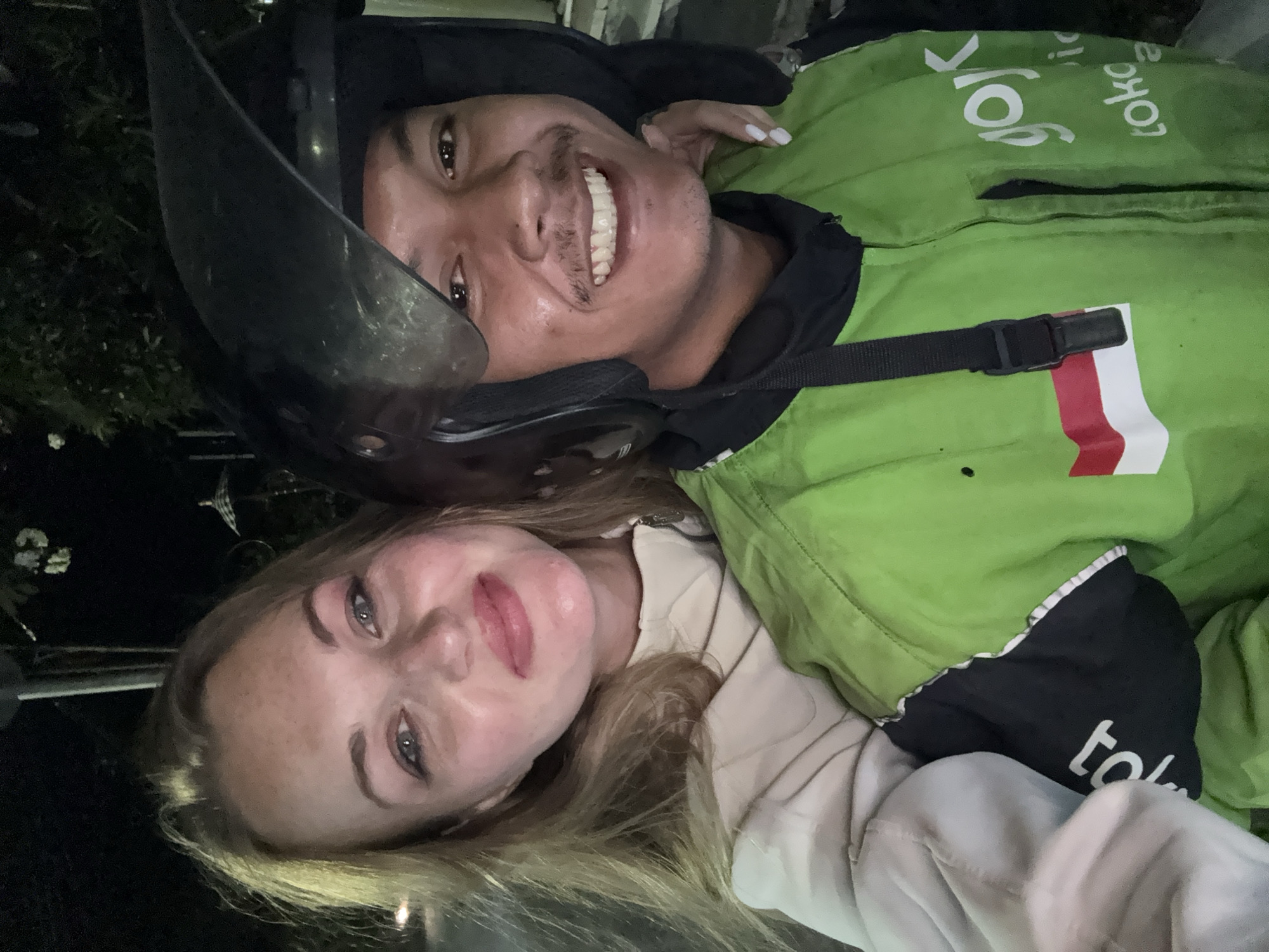 Two individuals are captured in a close-up, selfie-style photograph at night, likely in Denpasar, Indonesia. The person in the foreground on the right is a male wearing a black motorcycle helmet and a bright green jacket with the "Gojek" logo and an Indonesian flag patch. He is smiling, revealing white teeth. The person in the background on the left is a female with blonde hair, wearing a light-colored jacket. Her arm is visible around the male's shoulder, and her hand with light-colored fingernails rests on his jacket. The environment appears to be outdoors, with dark foliage and possibly streetlights or ambient lighting visible in the background. The overall impression is a casual, candid moment captured during a nighttime journey, possibly as passengers on a motorcycle.