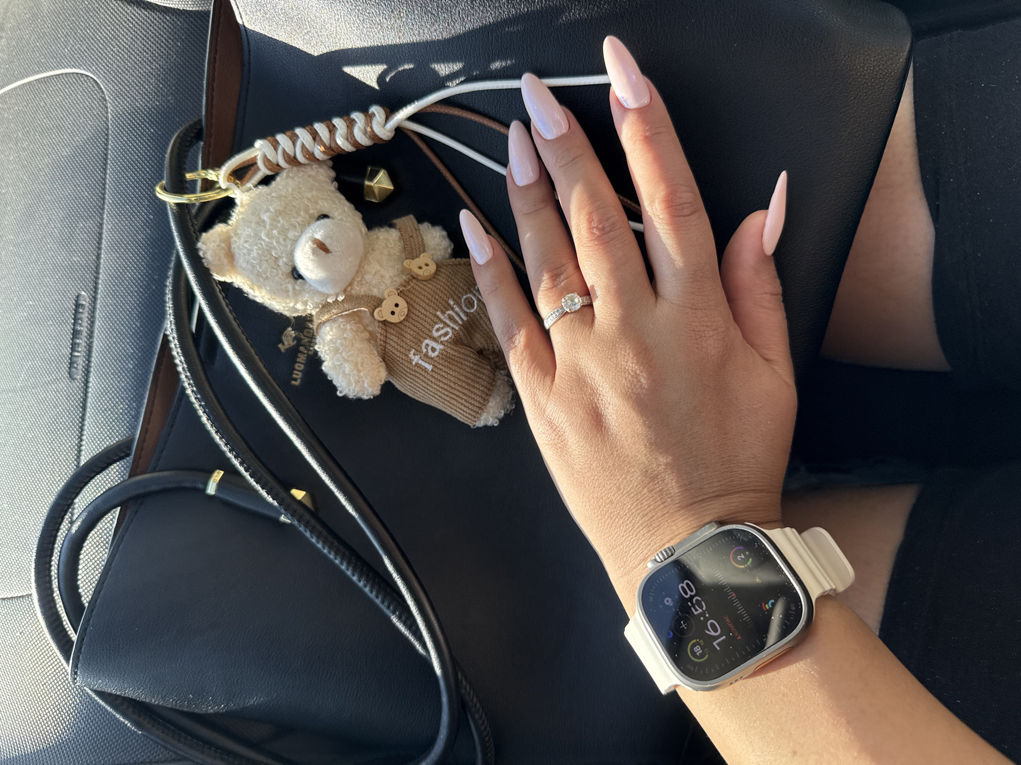 An overhead, close-up, eye-level shot captures a person's left hand and wrist, adorned with a white Apple Watch Ultra and a diamond ring, positioned over a dark leather handbag with a fluffy teddy bear charm attached. The watch face displays the time as 16:58 and several app icons. The hand has long, light pink almond-shaped nails and a diamond engagement ring on the ring finger. The handbag is dark, possibly black, with brown accents and a gold charm attachment. The teddy bear charm is small and light brown, wearing a tan overalls-like outfit with the word "fashion" written on it. The image is lit with natural light, casting shadows. The background appears to be the interior of a vehicle. There are no visible landmarks or signs that would indicate the location is in Tallinn, Estonia.