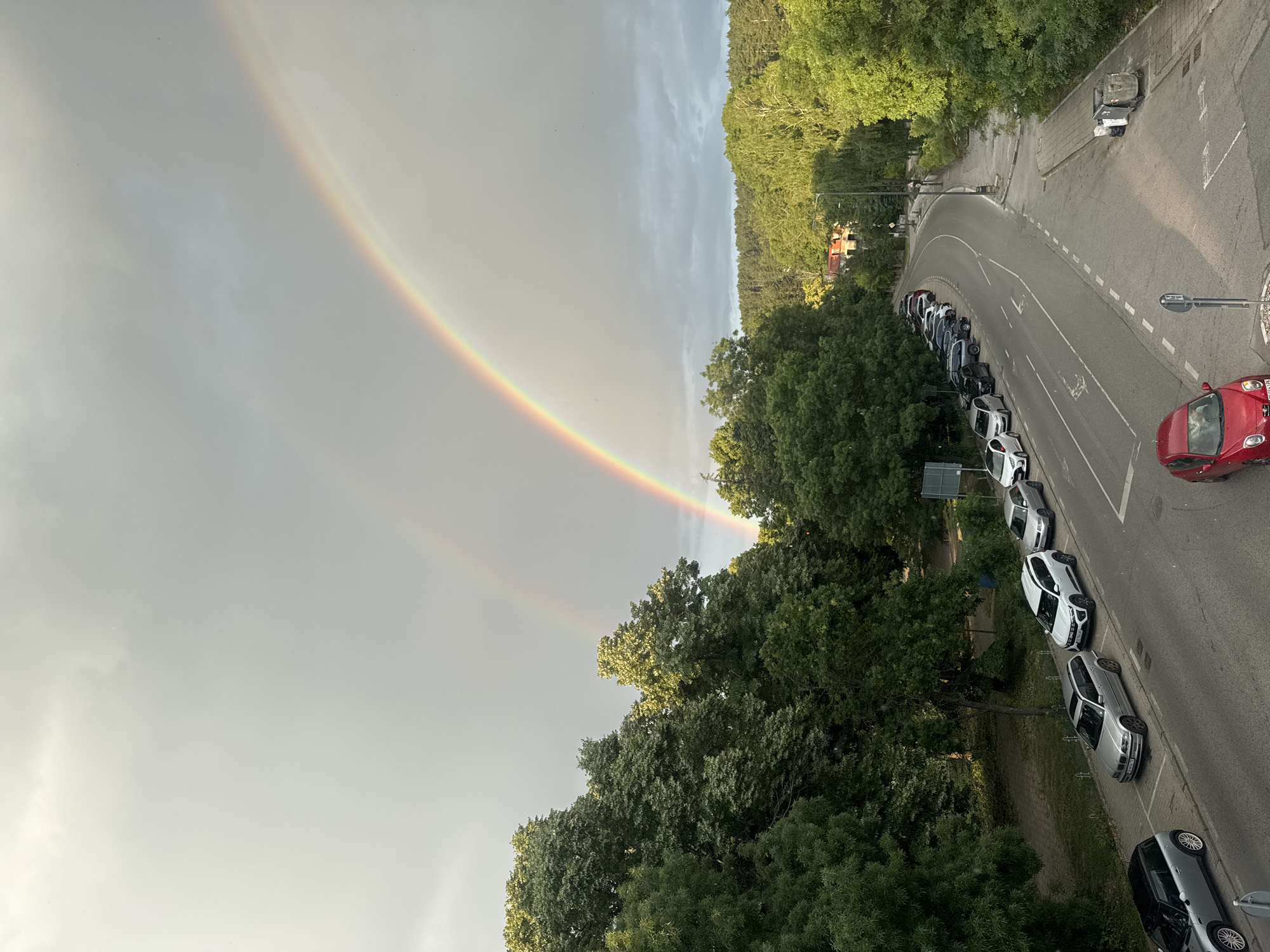 The image captures a double rainbow arching across a gray, cloudy sky, possibly after a rain shower. The scene is a street lined with parked cars, situated next to a dense forest of green trees. The perspective is from above, looking down at the street and cars. The cars are parked along the right side of the curved road. Some buildings with red roofs are visible nestled amongst the trees in the background. The time of day appears to be late afternoon or early evening, judging by the lighting and the presence of a rainbow. The overall mood is serene, with the natural beauty of the rainbow contrasting with the urban elements. No visible text is present in the image. The location is identified as Bankya, Bulgaria.