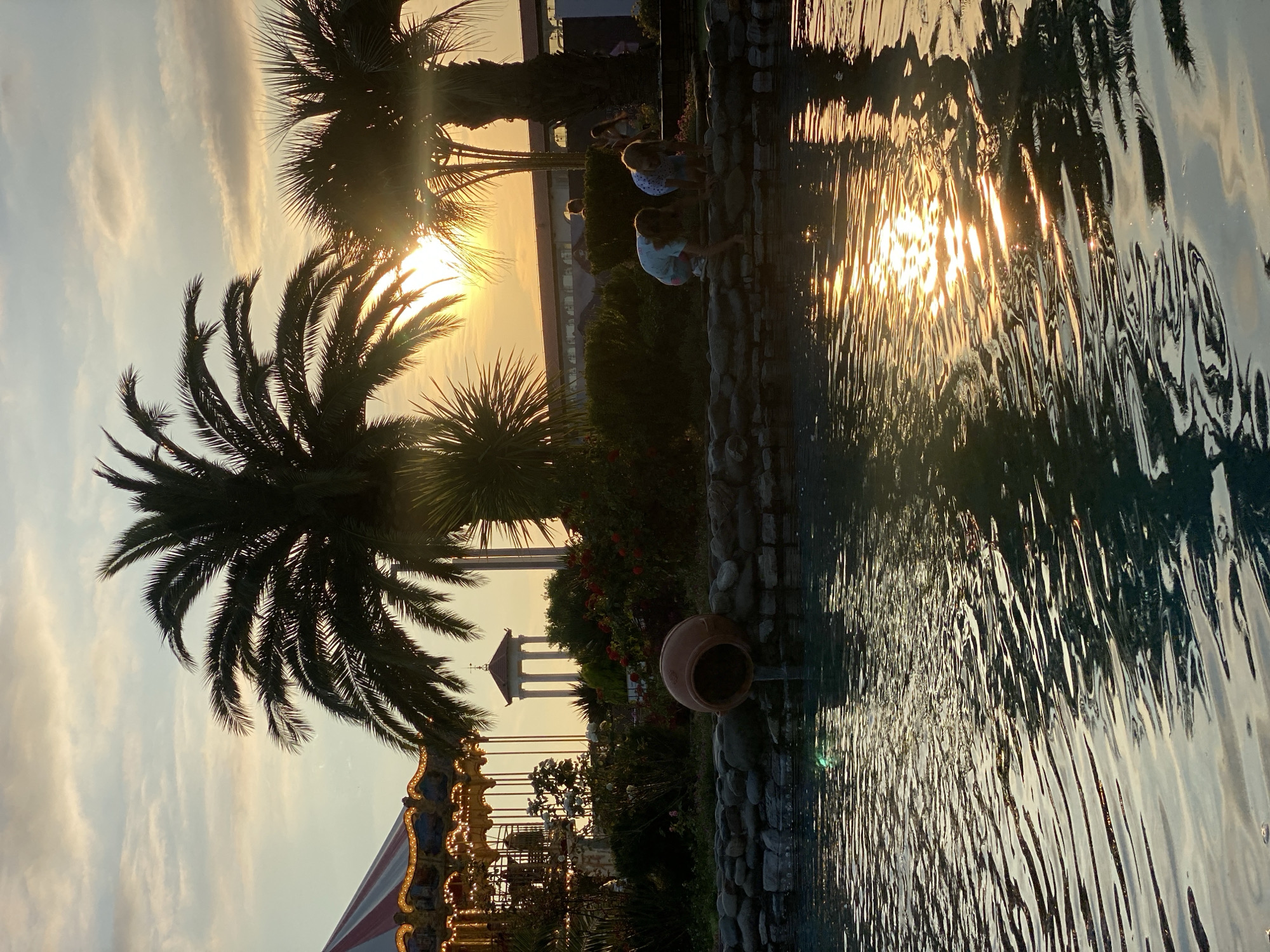 This image depicts a serene outdoor scene at sunset, likely in Sochi, Russia, given the context. The setting appears to be a park or recreational area, featuring lush palm trees silhouetted against a warm, golden sky. The sun is low on the horizon, casting a shimmering reflection on the water's surface, which is disturbed by ripples, creating an abstract play of light and shadow.
In the foreground, a body of water, possibly a fountain or a decorative pool, is bordered by stones. Several young individuals are gathered by the water's edge, their figures partially obscured by the low sunlight and reflections. They appear to be interacting with the water, perhaps dipping their hands or feet, suggesting a moment of casual enjoyment and relaxation.
Further in the background, a carousel with illuminated lights is visible, hinting at a carnival or amusement park atmosphere. Beyond that, a classical-style gazebo or structure with columns stands against the backdrop of the sunset. The overall mood is tranquil and peaceful, amplified by the warm hues of the sunset and the gentle movement of the water. The presence of palm trees and a carousel suggests a pleasant, possibly tropical or sub-tropical, tourist destination. The time of day is clearly sunset, characterized by the soft, diffused light and the golden glow across the sky. The weather appears clear and calm, conducive to outdoor activities. There is no visible text in the image.