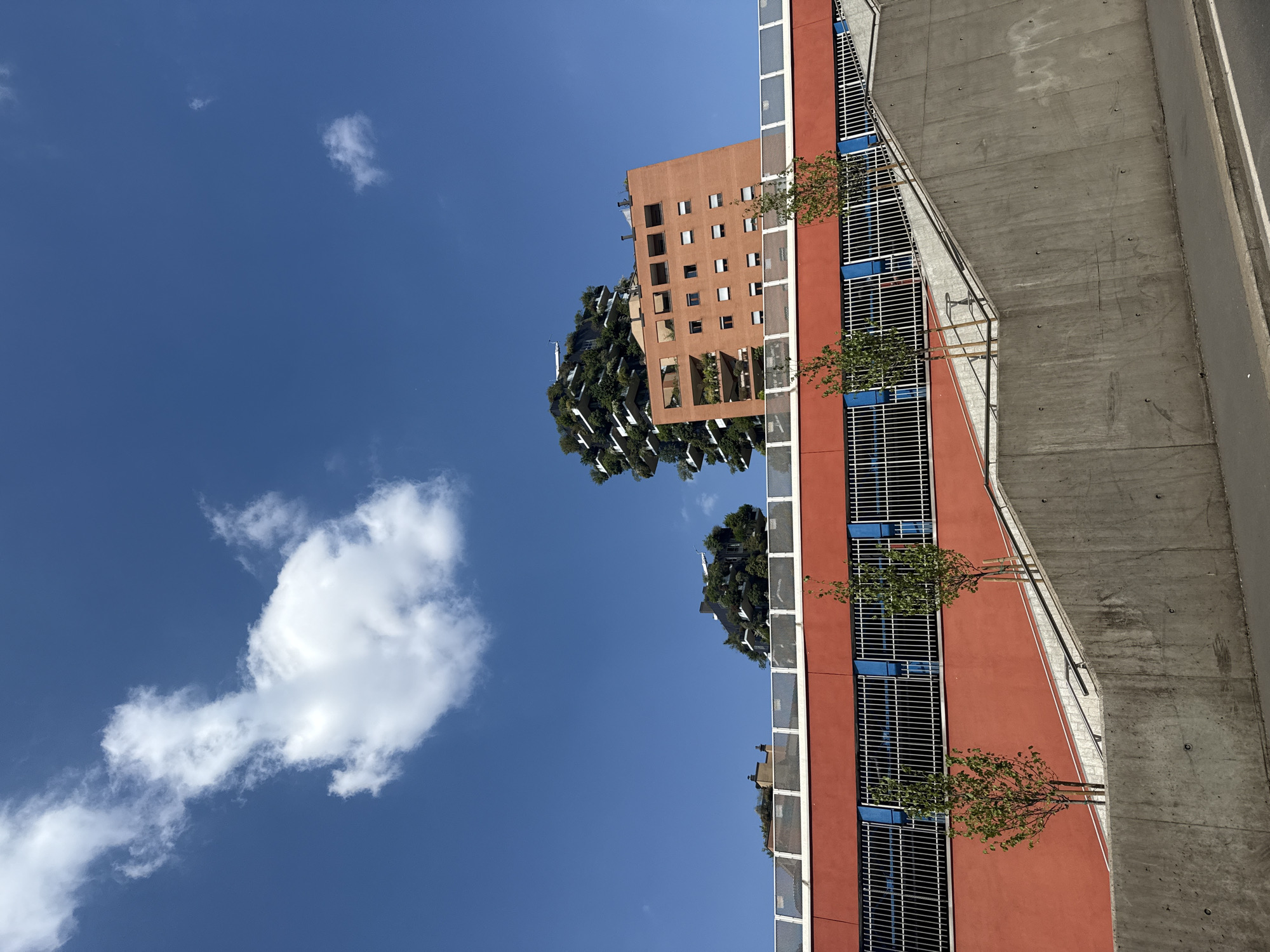 The image, when viewed upright, presents a modern urban scene under a bright, clear blue sky. In the foreground, a building with a reddish-terracotta facade occupies the lower right portion. This building features multiple horizontal white grates, possibly balconies or screens, interspersed with distinctive rectangular blue elements. Small trees or shrubs are planted in planters along its base.
To the right of this terracotta building, a wide concrete elevated structure or walkway with a metal railing is visible, leading to a darker asphalt road. The concrete surface shows some texture and subtle markings.
In the midground, set against the sky, are two other prominent buildings. The most striking is a "Bosco Verticale" (Vertical Forest) style building, characterized by numerous balconies and facades heavily covered with dense green trees and vegetation, giving it a lush, organic appearance. Behind and slightly to the right of this green building, a plainer, light brown or tan building with numerous square windows is visible.
The sky is a vibrant, deep blue, dotted with several scattered white, fluffy cumulus clouds of varying sizes. The strong, direct sunlight indicates a clear day, likely late morning or early afternoon. There are no people visible, and no discernible text appears in the image. The unique architecture, particularly the "Vertical Forest" building, serves as a strong location cue, indicating the scene is in Milan, Italy. The overall mood is serene, highlighting architectural innovation and the integration of nature within an urban environment.