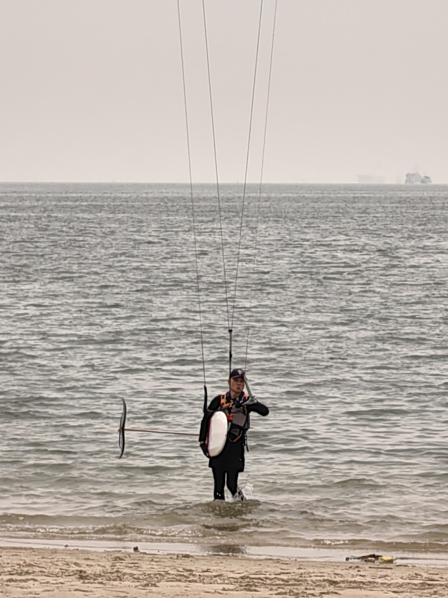 A man stands waist-deep in the water at Bai Chay Ward, Vietnam, carrying a hydrofoil board. He is wearing a black wetsuit and a safety vest. He is holding the hydrofoil with one arm and is holding up the kite lines with his other hand. In the background, the sea fades into a grey sky and a boat is visible far in the distance. The foreground consists of a sandy beach with some scattered debris.