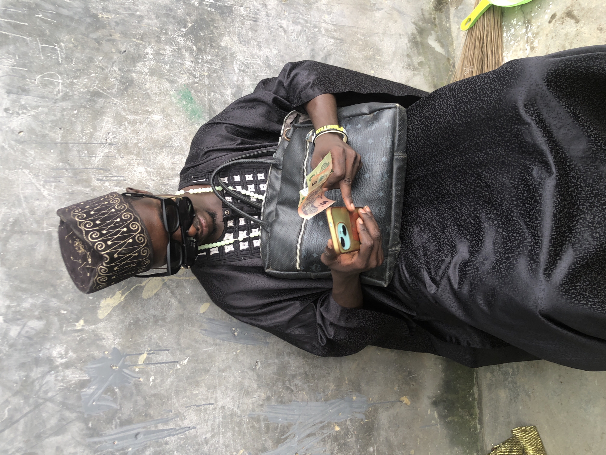 A man in traditional attire sits in front of a weathered concrete wall in Ado, Nigeria. He wears a black cap with gold embroidery, black sunglasses, and a black kaftan with a white and black patterned neckline. Around his neck is a beaded necklace, and he has a yellow and white bracelet on his left wrist. He is holding a black handbag with a monogram pattern and a cell phone in his hands, counting money. The ground appears to be concrete, and a broom is visible to the right of the man. The wall behind him has graffiti and paint splatters.