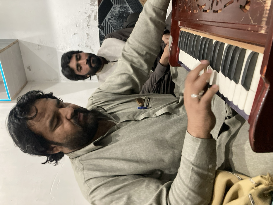 The image depicts two men indoors, seated in what appears to be a residential or informal gathering space in the city of Unknown, Unknown. In the foreground, a man with dark hair and a full beard, wearing a light grey long-sleeved traditional shirt, actively plays a brown wooden harmonium. His right hand is positioned on the instrument's white and black keys. A silver ring is visible on his right ring finger. A pen and a nametag reading "A-One" are clipped to his left shirt pocket. He is looking downwards, focusing on the harmonium. A beige fabric is visible on his left side. In the background, partially obscured, a second man with dark hair and a beard is seated, wearing a light-colored waistcoat over a dark shirt. He is looking forward. The background features light-colored walls, with the wall behind the second man showing peeling paint. A section of patterned, possibly upholstered, seating is visible behind the second man. A light-colored, shelf-like structure with a blue top border is mounted on the wall in the upper right.
