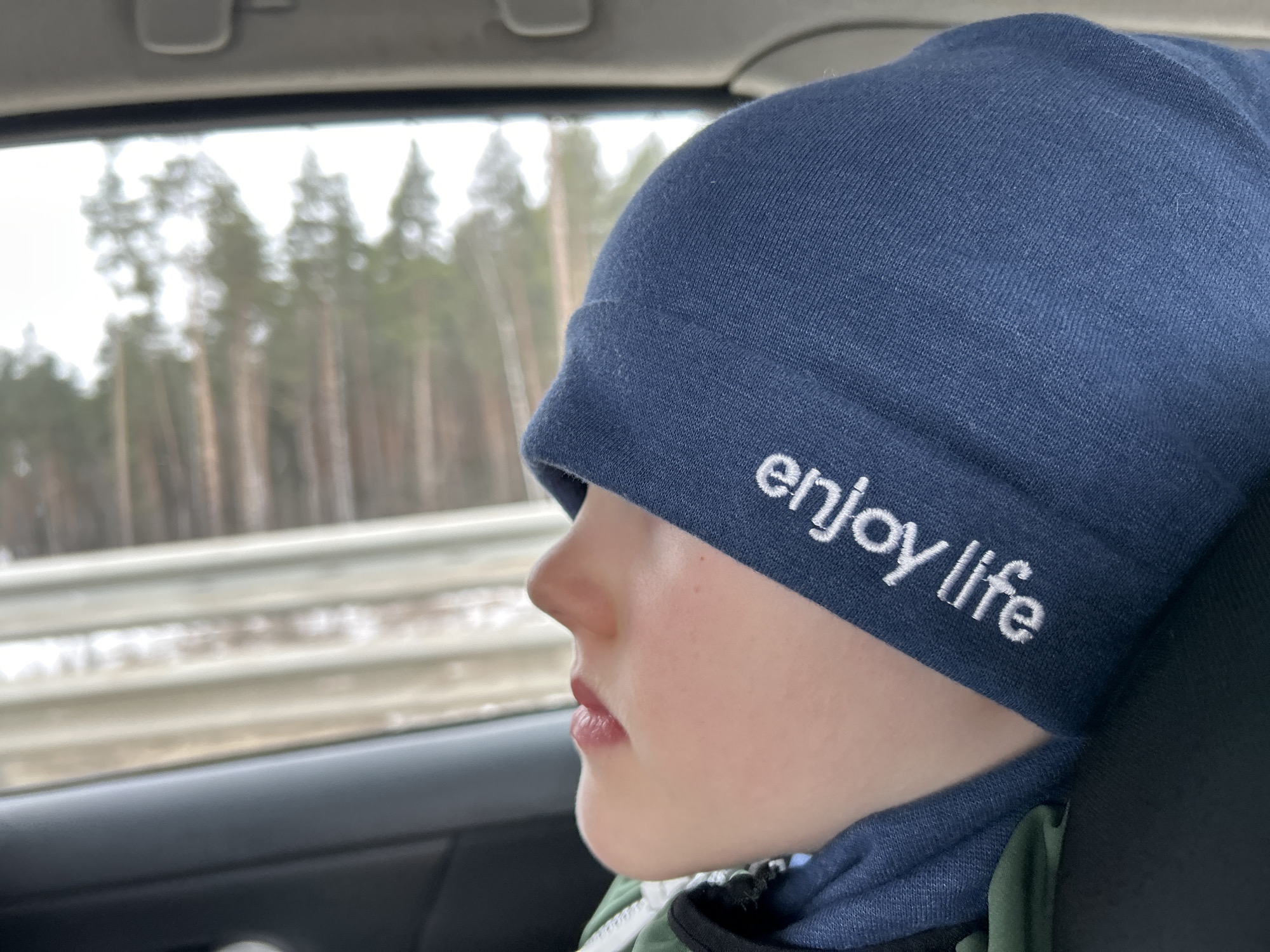 A young individual, appearing to be a child or pre-teen, is shown in a profile view, looking out the left-side window of a vehicle. The individual wears a dark blue knit beanie embroidered with the white text "enjoy life" and a dark blue neck gaiter or collar over what appears to be a green and white garment. Facial features visible include a light skin tone, a prominent nose, and slightly parted lips. The interior of the vehicle, including a dark door panel and window frame, is visible in the foreground. Outside the window, a blurred background depicts a winter or early spring landscape with tall, slender evergreen trees against an overcast sky. A metallic roadside guardrail is also partially visible through the window. This scene is located in Kosherovo, Russia.