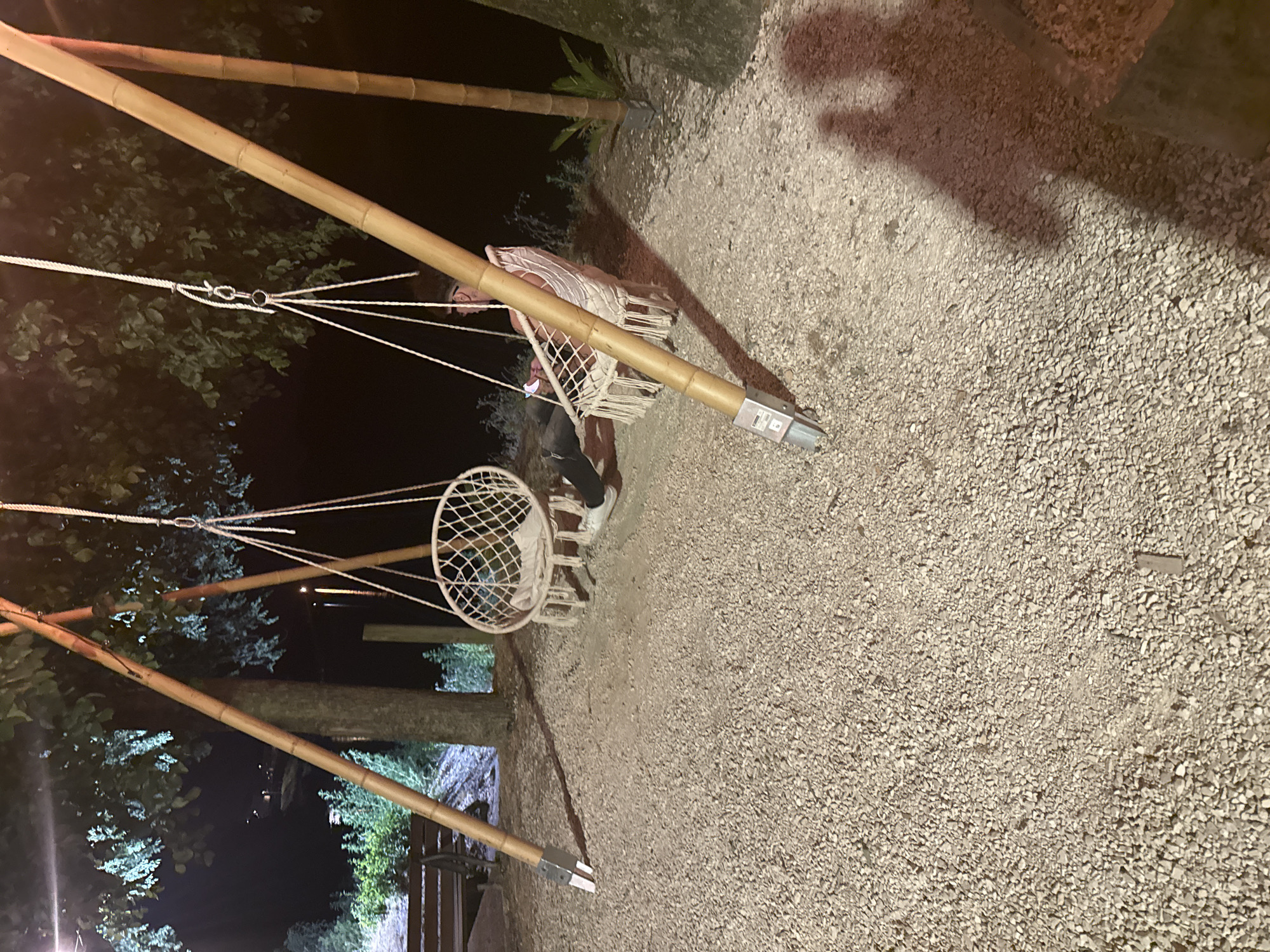 The scene depicts a person sitting on a swing, presumably outdoors and at night, supported by a bamboo-like structure with ropes. The ground is covered with gravel. The environment is dark, with glimpses of trees and foliage in the background. The location is Casalmaggiore, Italy.