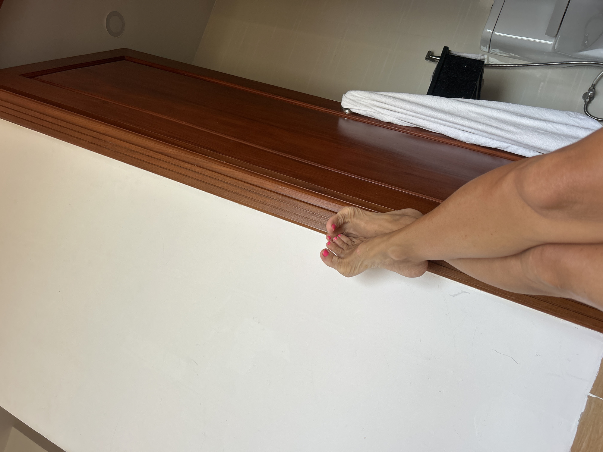 The image shows a portion of a bathroom interior, with white walls and a dark brown wooden countertop. The countertop appears to run under a white toilet. A white towel is present on the counter. A person's legs, with pink nail polish on the toes, are resting on the counter. The location is possibly in Bac Nha Trang Ward, Vietnam.