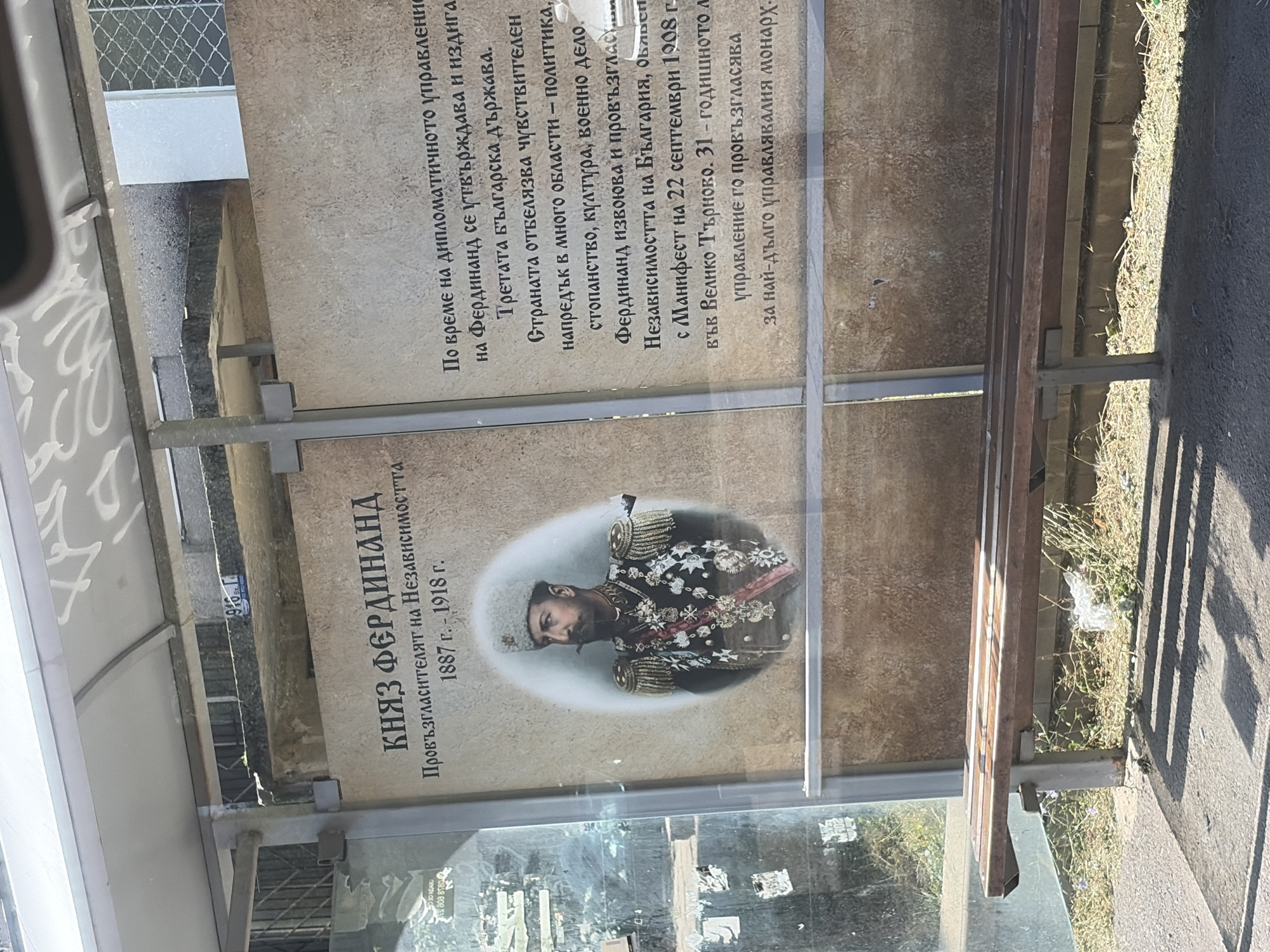 The image shows a bus stop with a large informative poster of Prince Ferdinand. The poster features a portrait of the prince in military attire and text in Bulgarian describing his reign. The bus stop has a wooden bench in the foreground and a concrete and metal structure. The environment appears to be outdoors, with sunlight casting shadows on the ground. There is some vegetation growing around the bus stop.