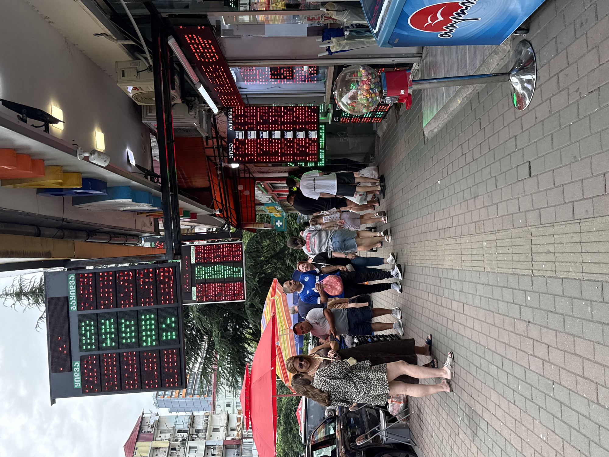 The image captures a street scene in an urban environment, possibly a commercial area, with several people interacting on a paved sidewalk. A large electronic display board, likely showing currency exchange rates, is prominently visible, with rows of red and green lights indicating values. Text in a foreign script appears above and within the display. Multiple people are standing and walking, some in casual attire. To the left, red and striped umbrellas provide shade. A black vehicle is parked nearby. Further down the sidewalk, a gumball machine stands on a metal pole. The buildings in the background appear to be multi-story structures. The overall atmosphere suggests a busy street during daytime.