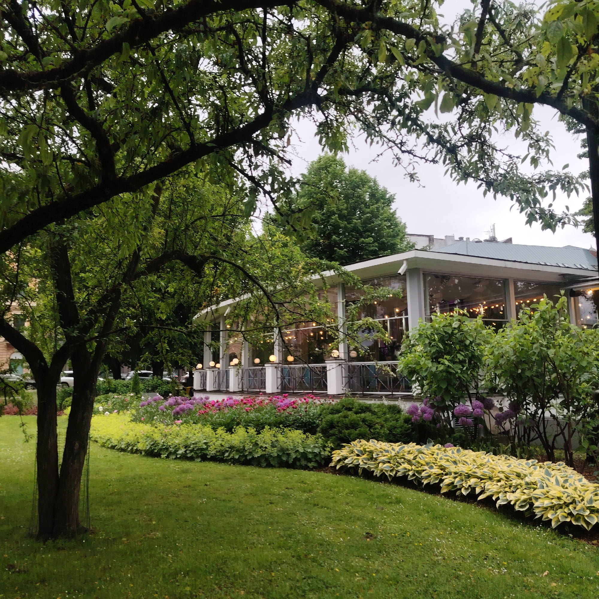 The image depicts a picturesque garden scene in front of a building with large glass windows, suggestive of a restaurant or cafe. The garden is lush with green grass, a variety of flowering plants in shades of pink, purple, and yellow, and mature trees providing shade. String lights are visible through the windows, indicating it may be evening or a dimly lit interior. In the background, some cars are parked along a street, hinting at an urban setting. The architecture of the building appears modern with white accents and a light-colored roof. The overall impression is one of tranquility and natural beauty within a city environment. This scene is located at the restaurant "Aqua Luna" in Riga, Latvia, known for its scenic garden and terrace.