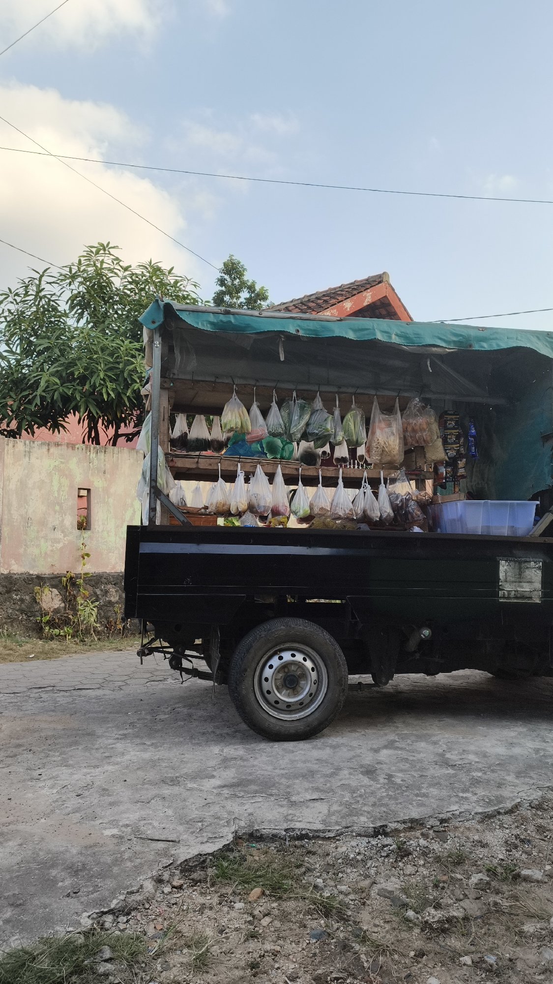 A mobile market stall, loaded with various goods, is parked on a street in Bandar Lampung, Indonesia. The stall is set up on the back of a black pickup truck, covered by a green tarp. Bags of produce, including what appear to be vegetables and fruits, are hanging from hooks above the truck bed. Shelves beneath the hanging bags display more packaged goods. A clear plastic container sits on the right side of the truck bed. The truck is parked on a concrete road with visible cracks and patches of gravel and grass. In the background, a weathered pink wall and a house with a red tile roof are visible, along with some trees and a cloudy sky.