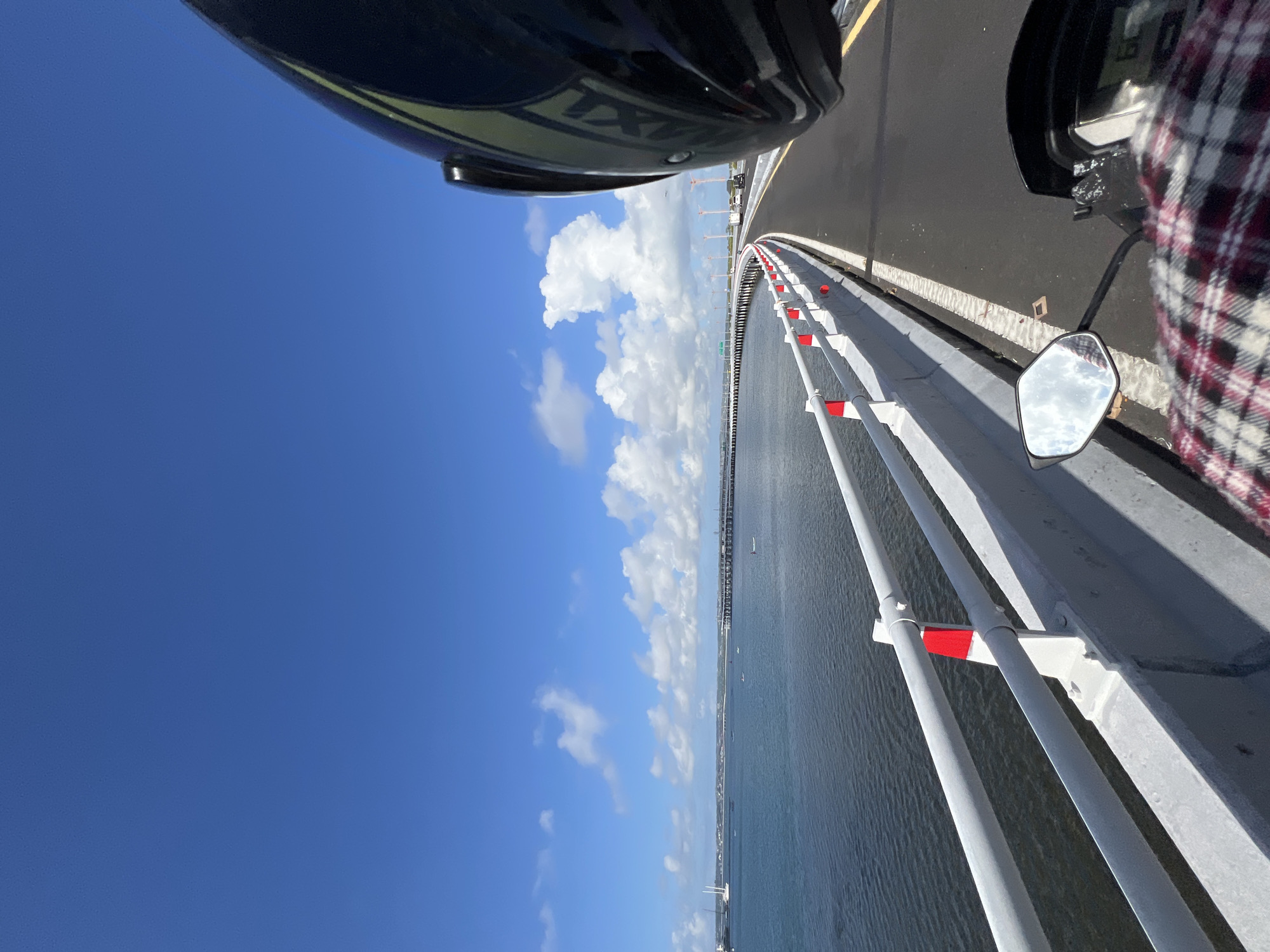 The image shows a view from a motorcycle while traveling on a bridge. The bridge has a concrete barrier with white railings, punctuated with red reflective markers. A body of water stretches out on the left, with a cityscape visible in the distance. The sky is blue with puffy white clouds. A motorcyclist's helmet is visible at the top of the frame, and a mirror of the motorcycle is reflecting the sky. The image suggests the location is coastal, with modern infrastructure.