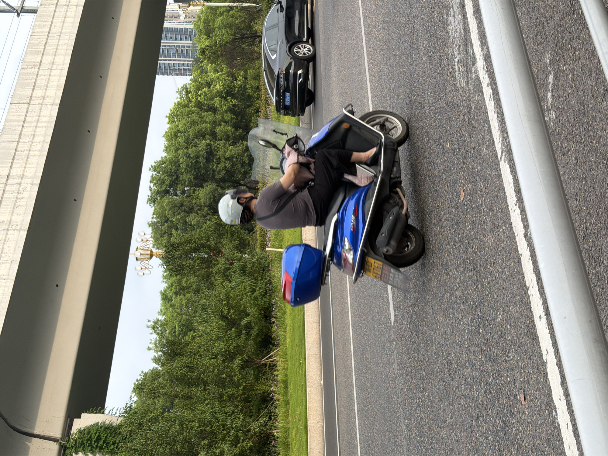 A person wearing a light-colored helmet and a gray t-shirt is riding a blue scooter on an asphalt road. The scooter has a red and blue storage box on the back. The scooter's license plate is visible and reads "6V767". A black sedan is driving in the lane next to the scooter. In the background, there are green trees, a streetlamp, and tall buildings. An overpass runs overhead. The scene appears to be in an urban area of Jiangjin, China.