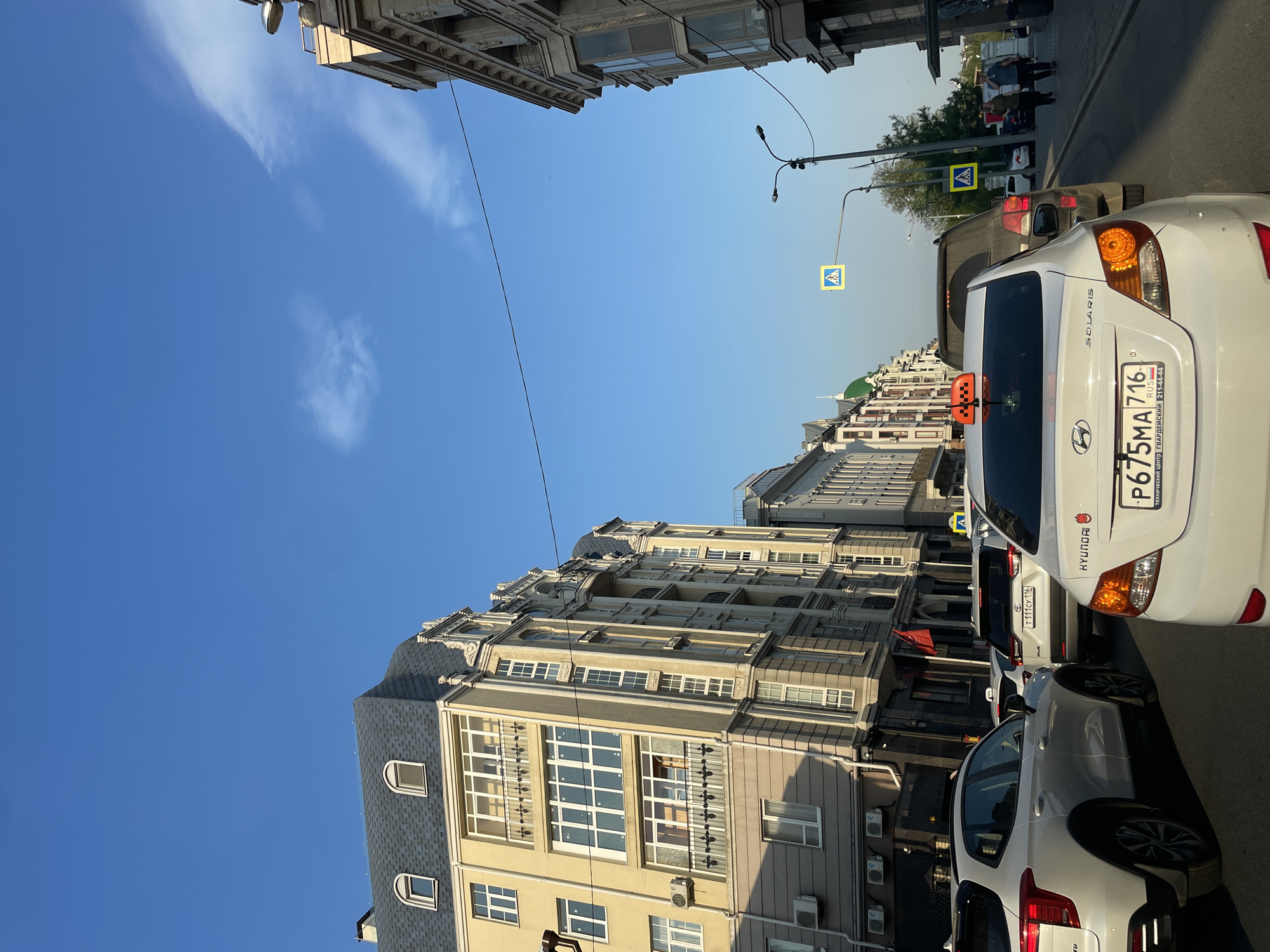 The image depicts a street scene in Kazan, Russia. A white Hyundai Solaris taxi with license plate P675MA716 is prominently featured, partially blocking the view of the street and buildings. Streetlights and pedestrian crossing signs are visible. Other vehicles are parked along the street, and several people are walking in the background. The buildings are multi-story structures with classical architectural details, typical of urban areas. The sky is blue with sparse clouds.