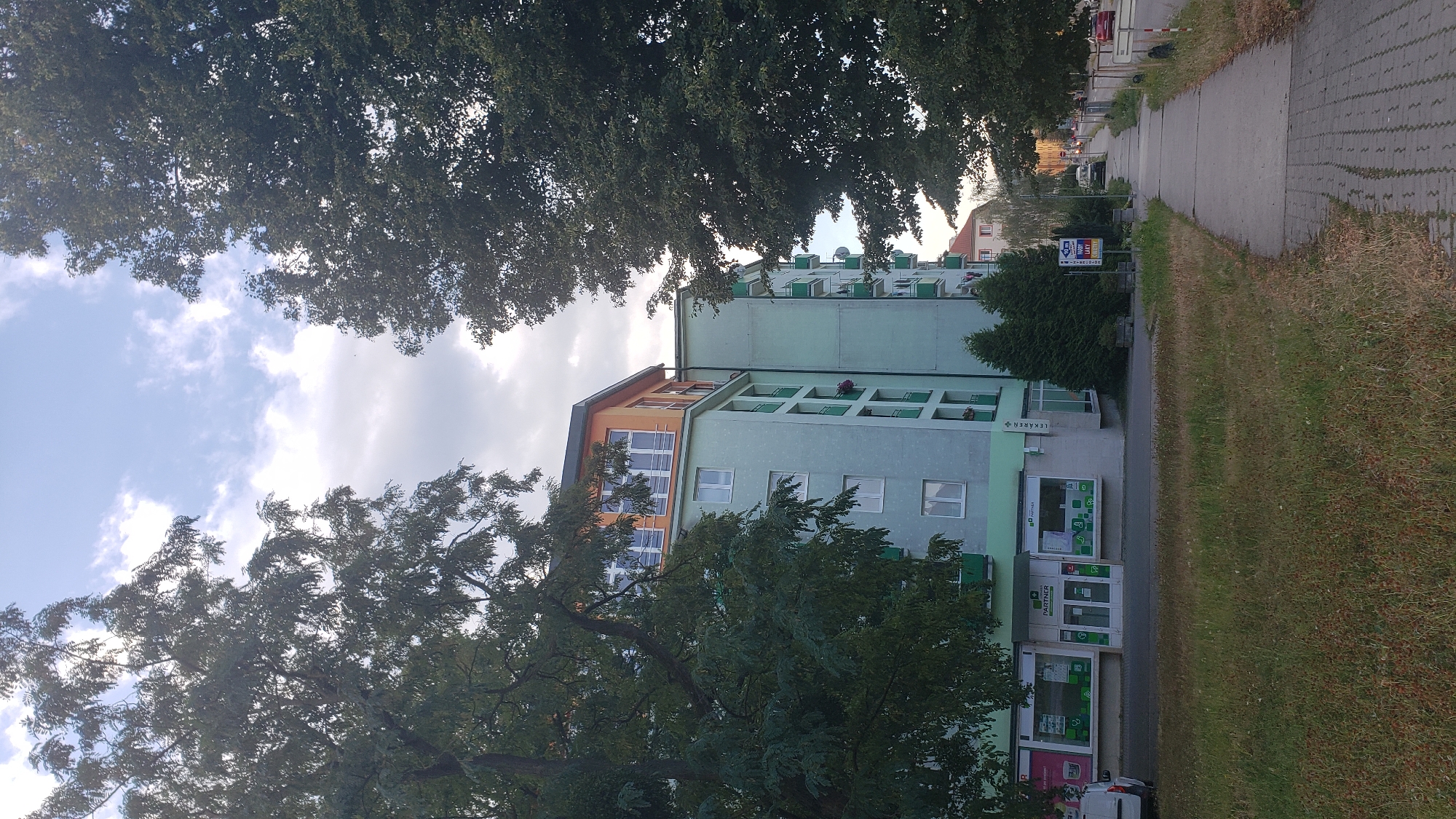 The image captures a street scene in Prešov, Slovakia, featuring a multi-story apartment building with a light green facade and an orange-colored top floor. The building is flanked by large, leafy trees, one on the left partially obscuring the view and another on the right framing the sidewalk. A sidewalk made of paving stones runs along the right side of the image, bordered by a strip of grass and then a road with a few parked cars. At the base of the apartment building, a row of shops is visible, including a "PARTNER" store with glass doors and display windows. The sky above is partly cloudy, with a mix of white clouds and patches of blue. The overall impression is of a residential area in a European city, possibly during daylight hours given the illumination.
