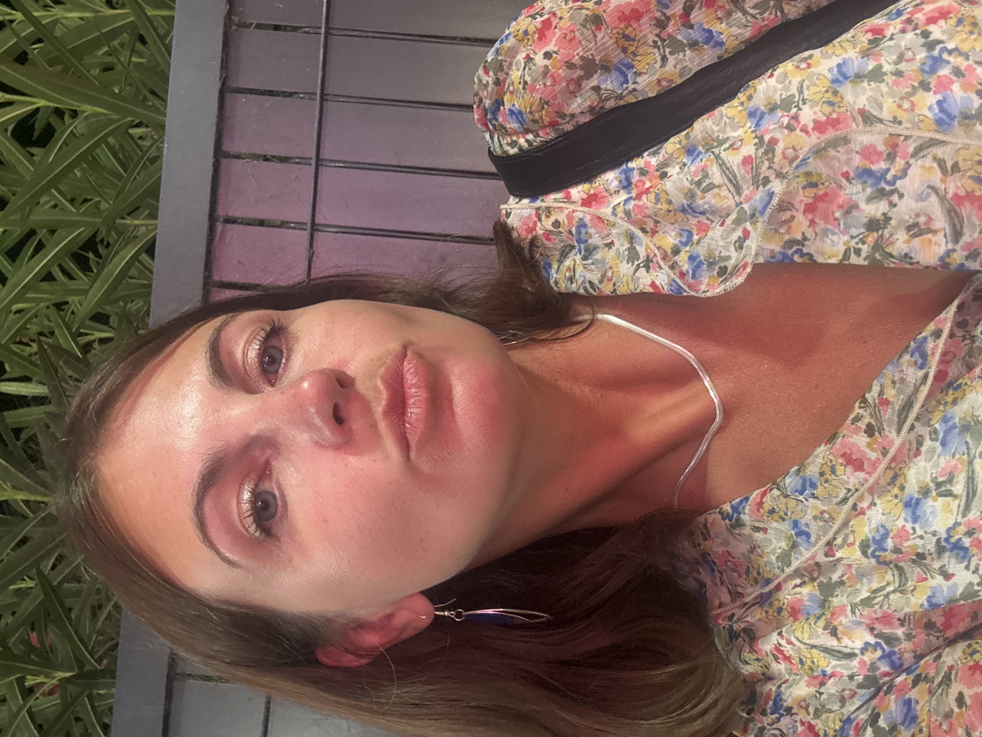 A woman with brown hair and blue eyes is taking a selfie. She is wearing a floral print dress and a silver necklace. She is also wearing a silver earring in her left ear. The background is blurred and appears to be outdoors. The scene takes place in Argelès-sur-Mer, France.