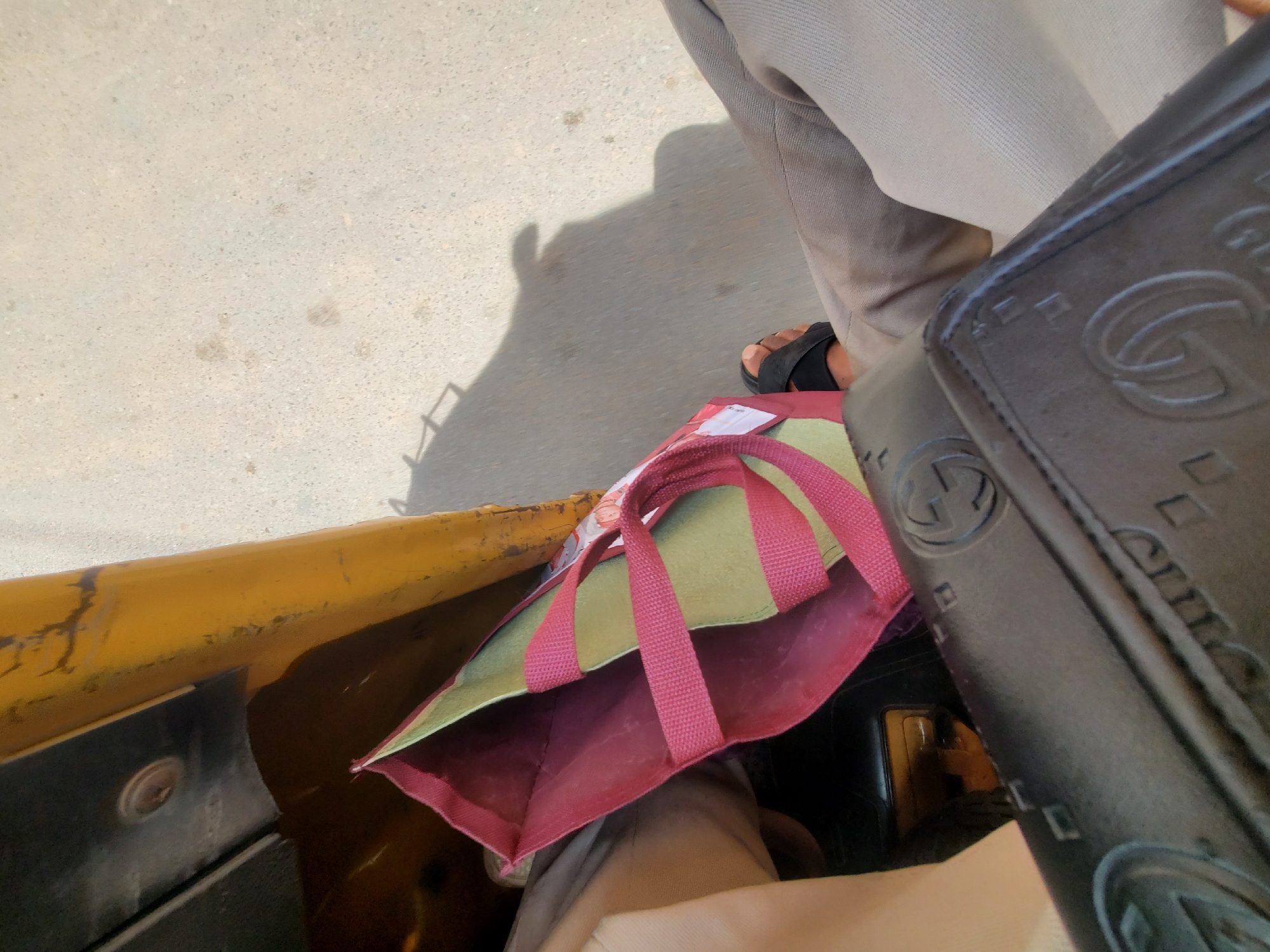 The scene is a close-up, angled overhead view, likely taken from the interior of an auto-rickshaw (keke) in Nkpor, Nigeria. Key elements include:
**People:** Partially visible, one person is wearing light brown trousers and black sandals.
**Subjects:** A reusable shopping bag with green and red panels and pink straps is placed on the floor of the keke, near the person's feet.
**Objects:** The interior of the keke, including the yellow frame and black paneling, is visible. A black object with repeating "G" logos is also present, obscuring part of the view. The street or ground is visible outside the keke.
**Environment:** The location is likely Nkpor, Nigeria, based on the prompt. The scene appears to be taken during daylight hours, given the bright lighting. The environment outside the keke appears to be a street or paved area.