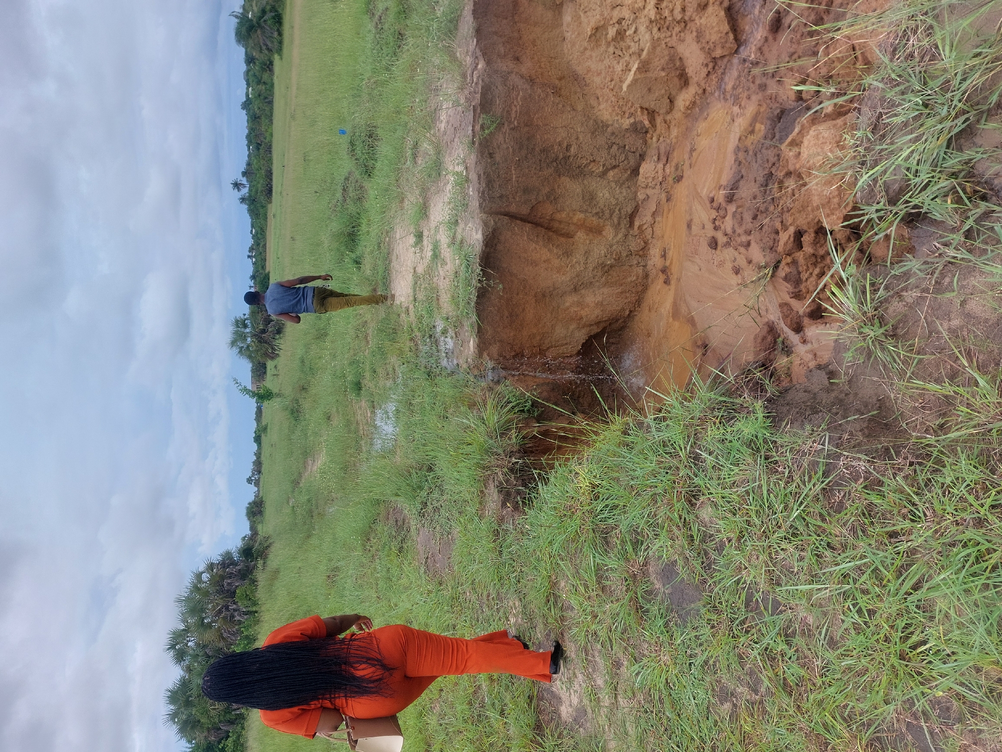 The image shows a scene in an outdoor location, likely in a rural or semi-rural area near Dar es-Salaam, Tanzania. There is a large erosion gully in the foreground, with exposed soil. Water appears to be flowing from the gully. Two individuals are present: one, wearing an orange outfit, is in the foreground, while another, wearing a gray shirt and brown pants, is standing farther away, closer to the erosion site. The surrounding environment features grassy vegetation. The sky is visible with scattered clouds.