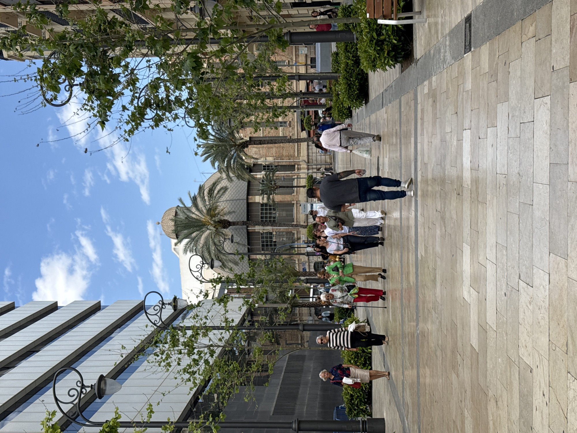 The image shows a wide, paved pedestrian walkway in an urban setting. A group of people is walking on the walkway, in front of a building with reflective, slanted panels. The walkway is lined with young trees and ornate street lamps. In the background, there is a building with a domed roof and palm trees. The sky is visible and mostly clear with some clouds. The location is in Unknown, Unknown.